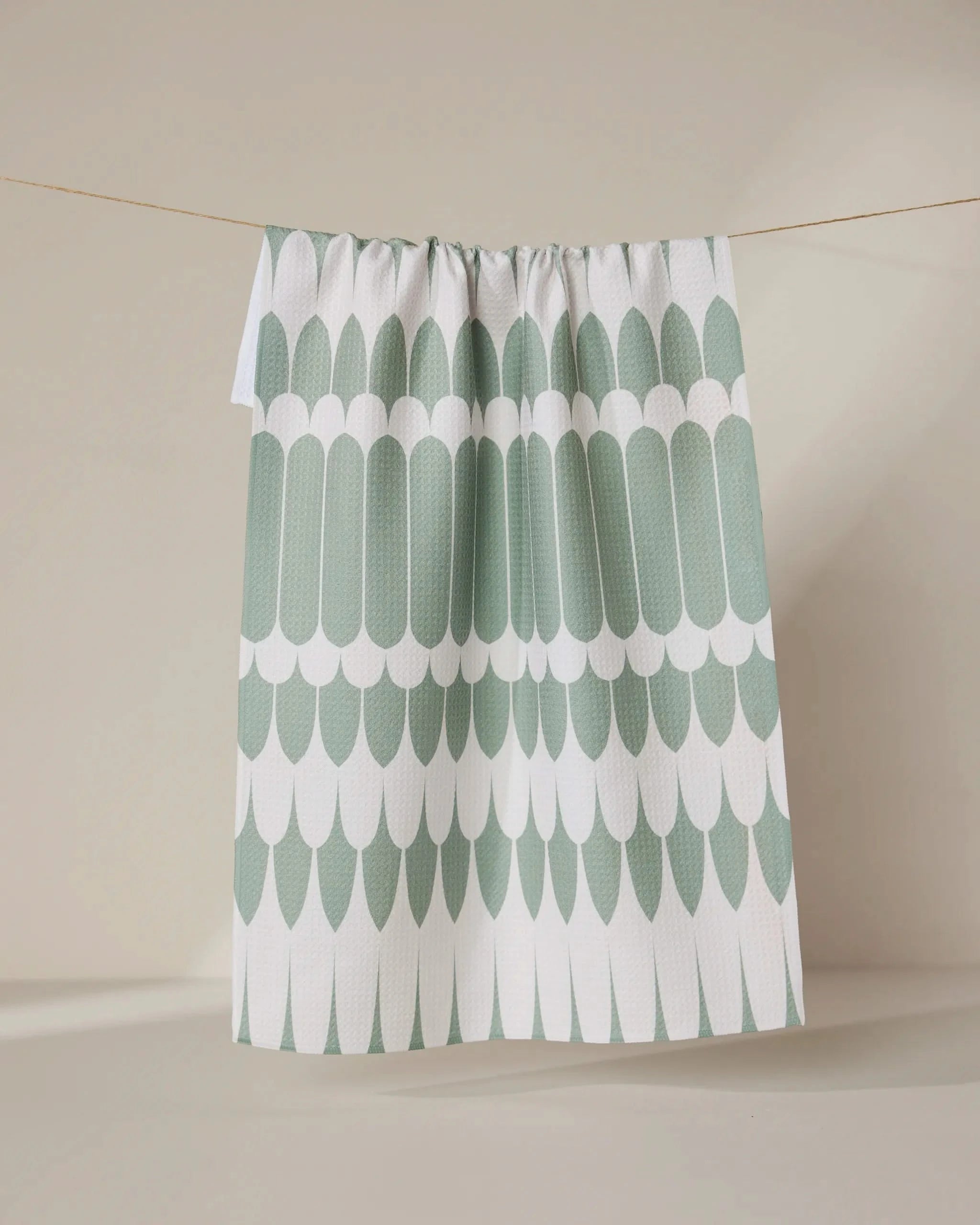 The GEOMETRY Salle Verte tea towel, featuring a green and white scalloped pattern, hangs on a clothesline against a neutral beige background.