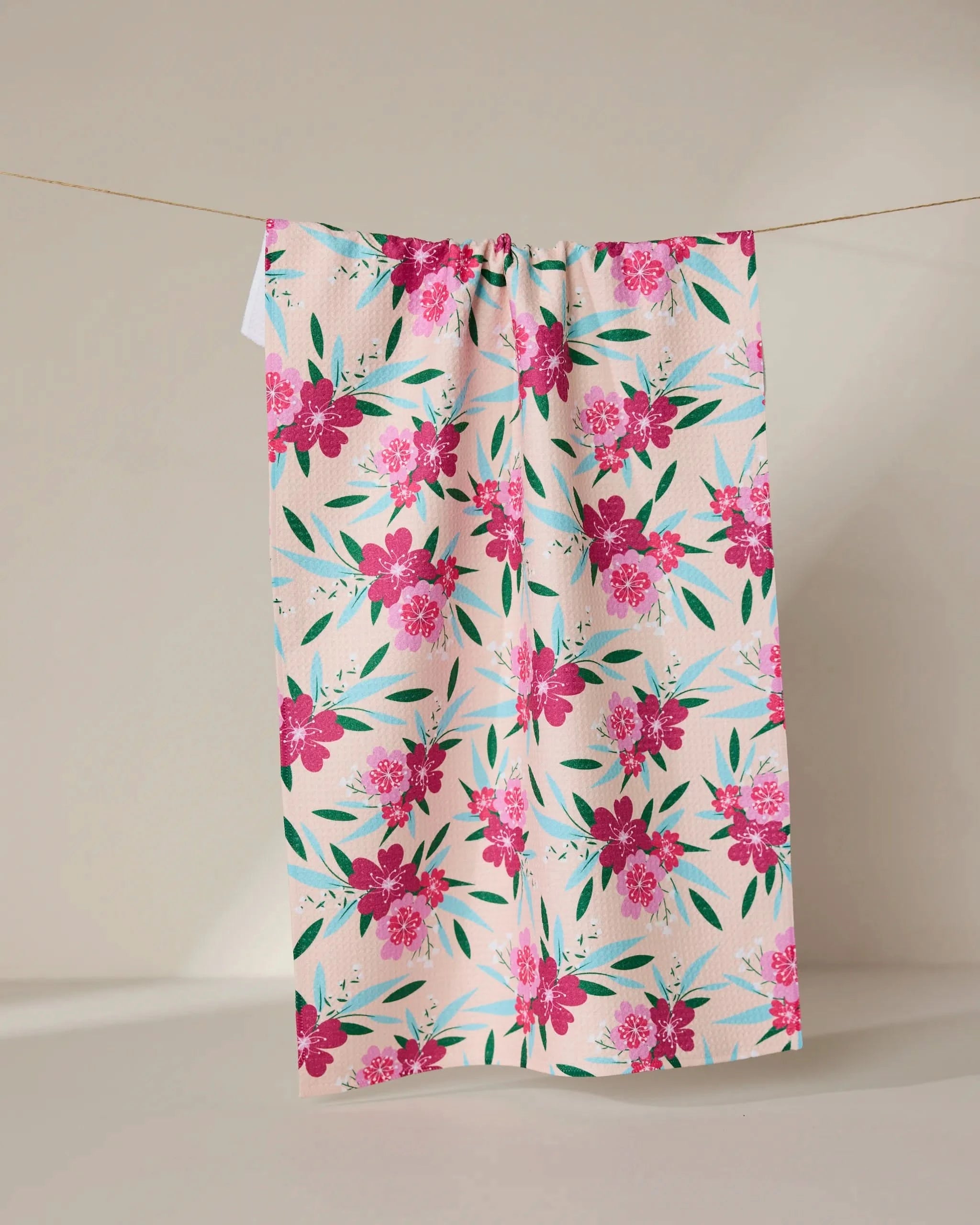The GEOMETRY Sending Love tea towel, crafted from recycled materials and featuring pink flowers with green leaves, hangs on a clothesline against a plain backdrop.