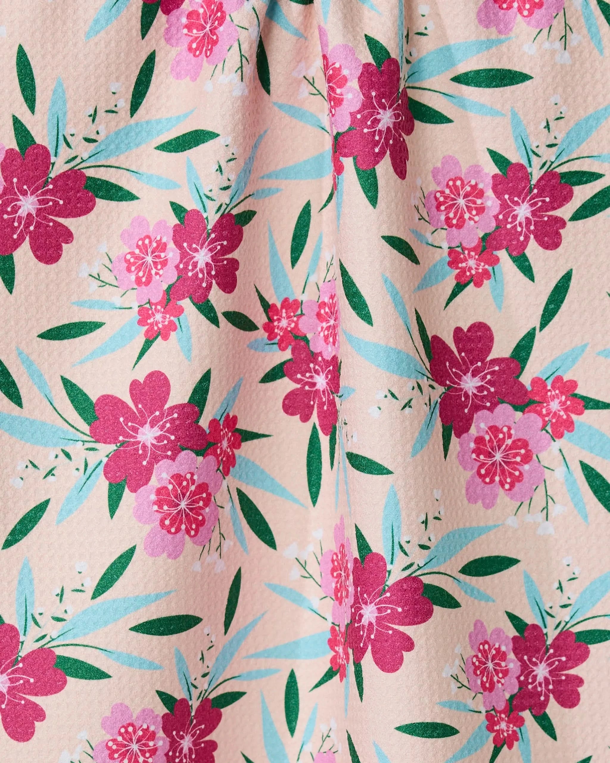 A close-up of GEOMETRYs Sending Love tea towel, made from recycled materials, features red and pink flowers, green leaves, and blue-green foliage on a pink background. The textured fabric is slightly gathered at the top.