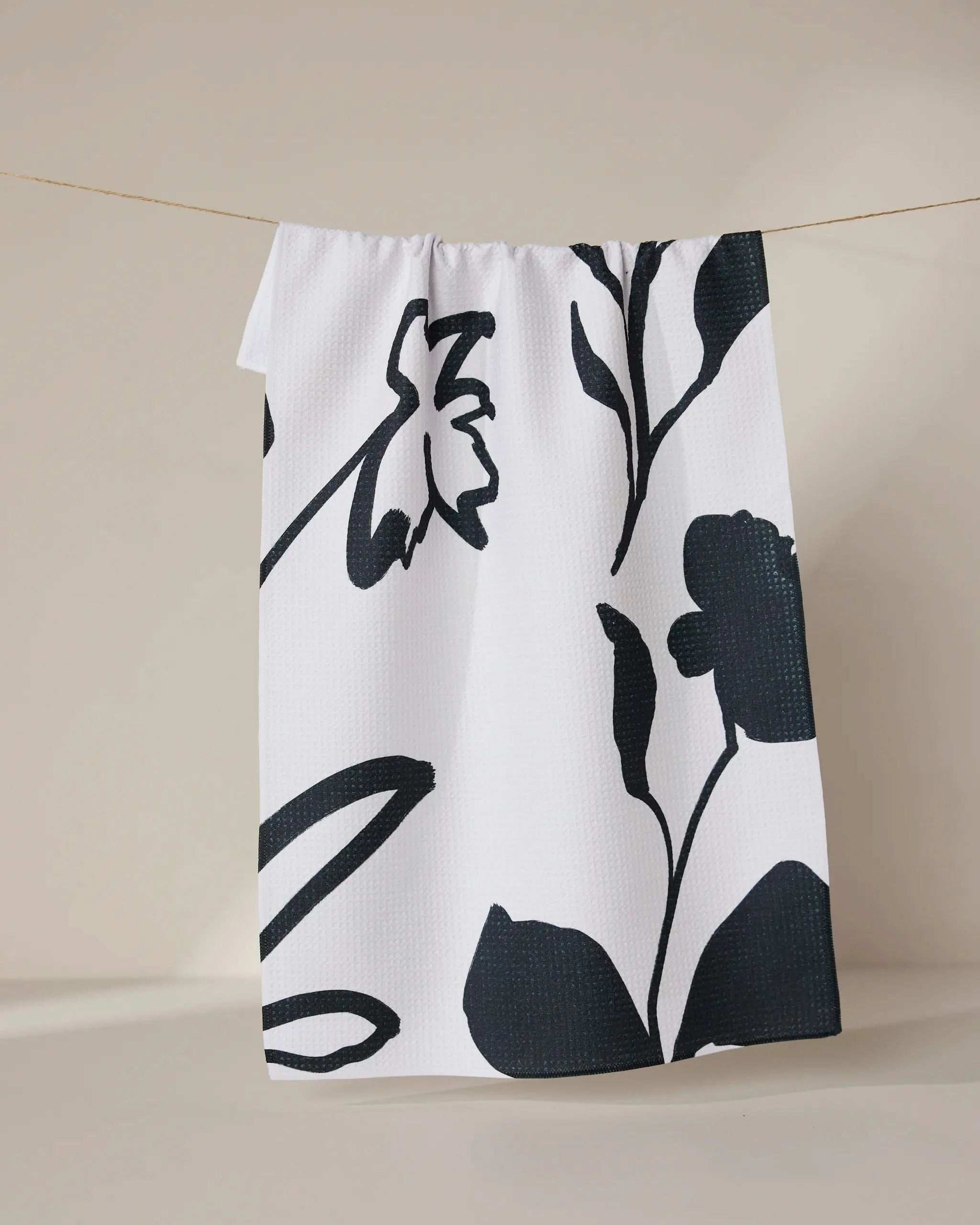 The Shade tea towel by GEOMETRY, featuring large bold black floral prints and made from recycled materials, hangs from a thin string against a neutral background.