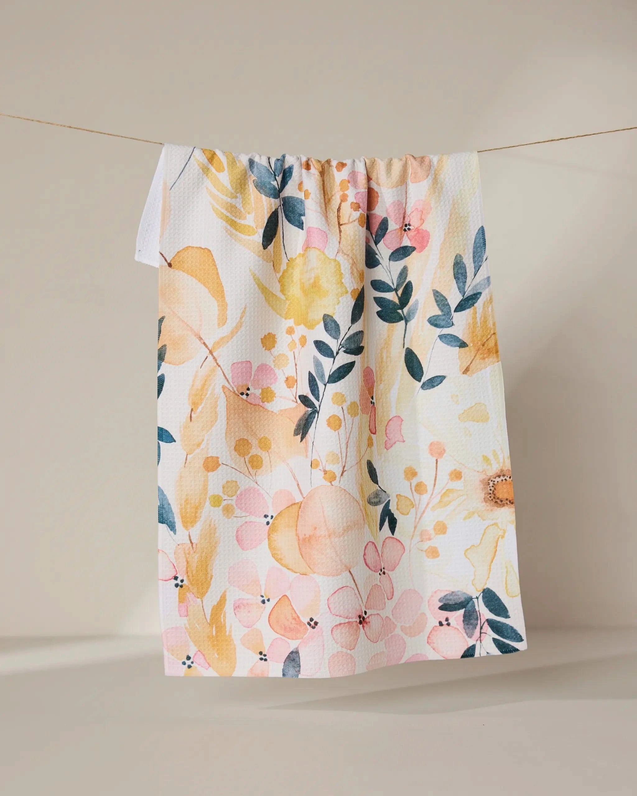 The GEOMETRY Sleepy Fields white kitchen towel, featuring a vibrant floral pattern in yellow, pink, and blue and made from recycled materials, hangs on a string against a neutral beige background.