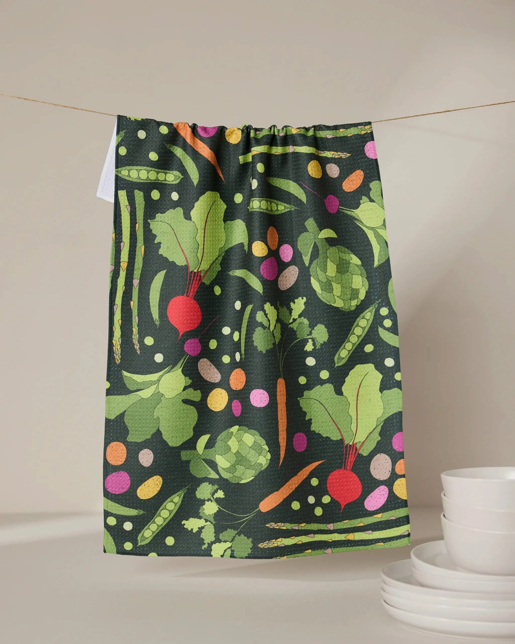 The GEOMETRY Spring Sprout tea towel, featuring a vibrant veggie pattern—beets, peas, artichokes, carrots, and leafy greens—crafted from recycled materials, hangs above stacked white bowls and plates against a neutral backdrop.