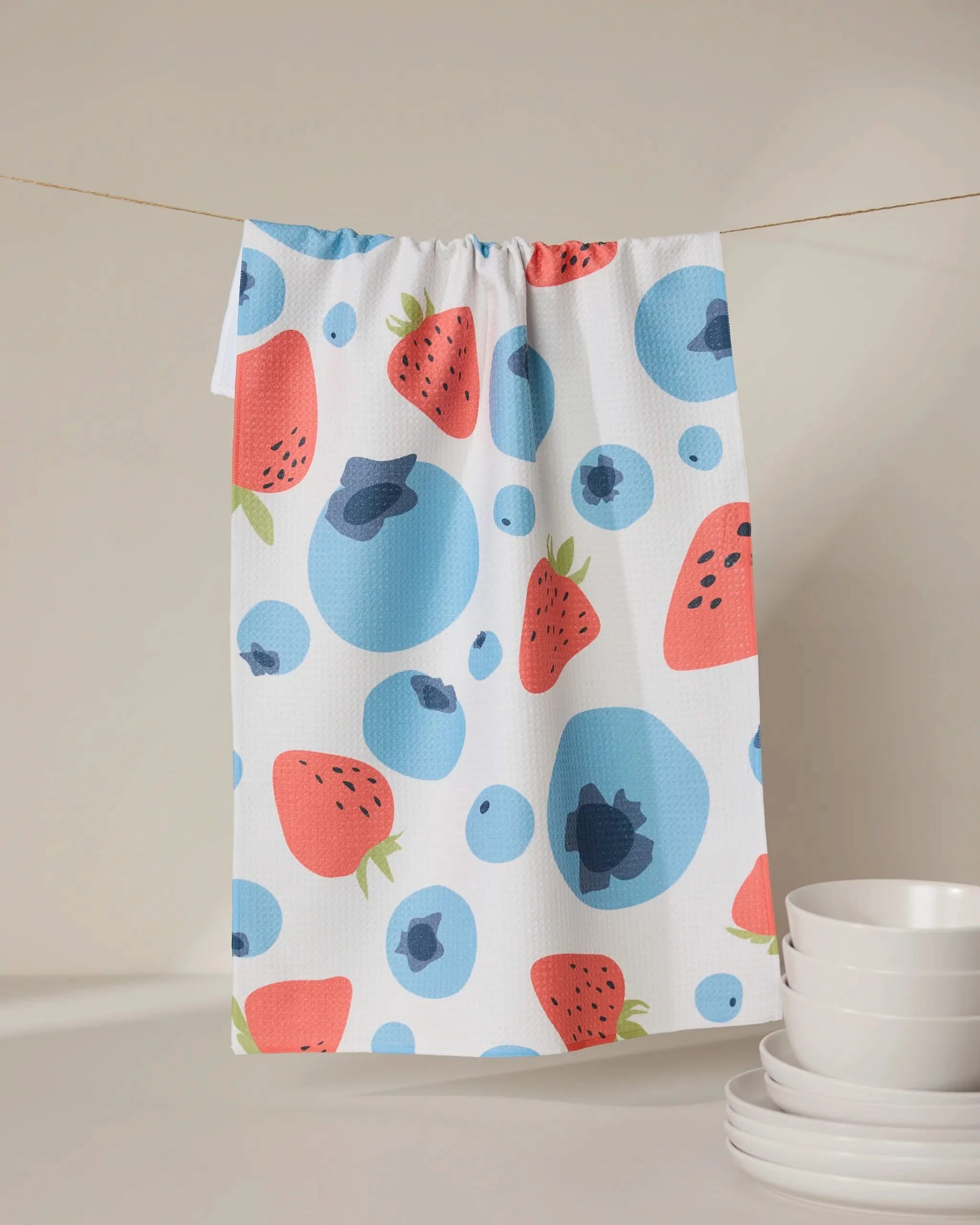 A GEOMETRY Sunny Berries recycled kitchen towel, featuring a vibrant strawberry and blueberry pattern, hangs on a string above stacked white bowls.