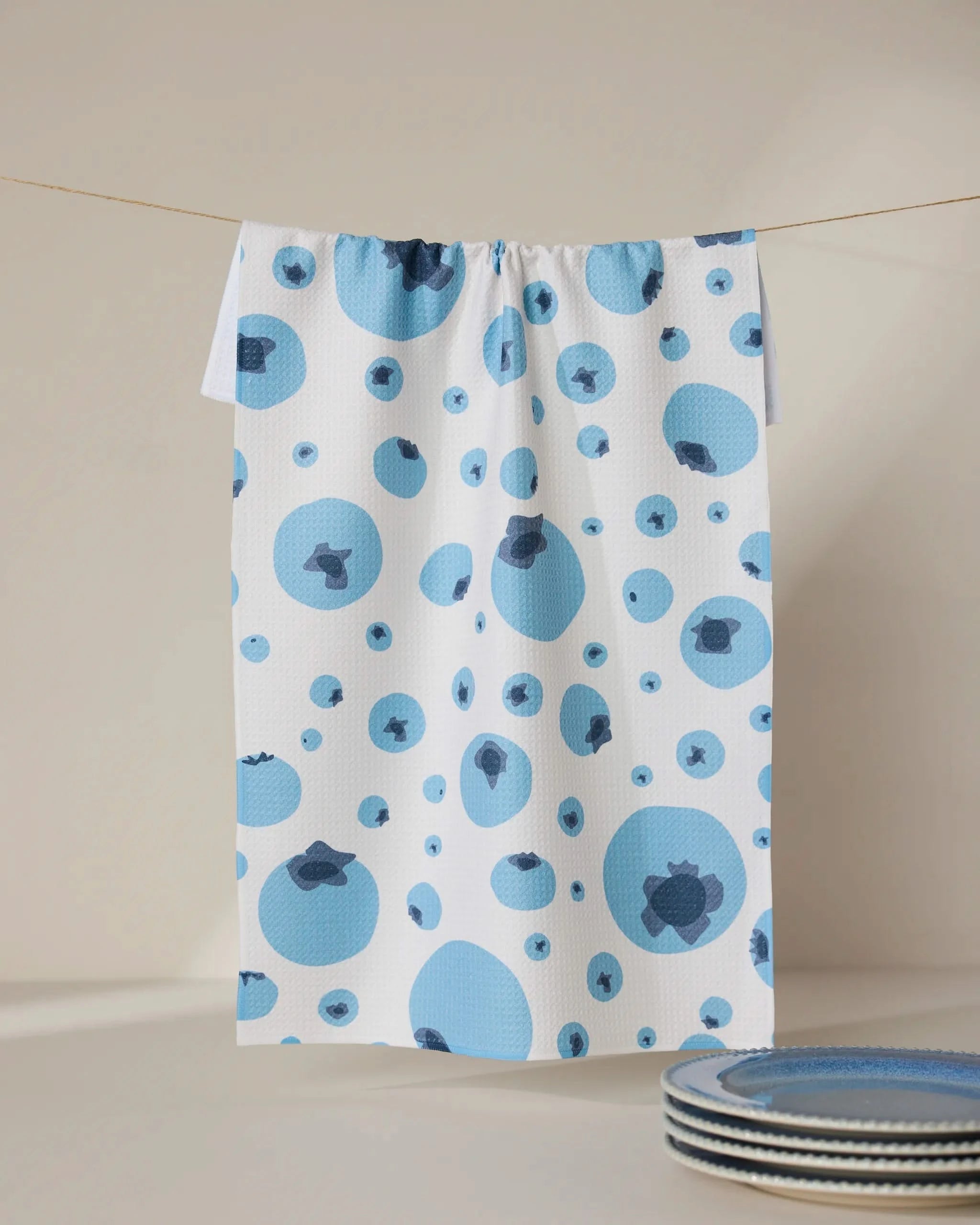 The GEOMETRY Sunny Blueberries white tea towel, crafted from absorbent Geoweave fabric and featuring a blue and dark blue blueberry pattern, hangs on a thin line. Blue-rimmed plates are partially seen in the lower right against a neutral background.