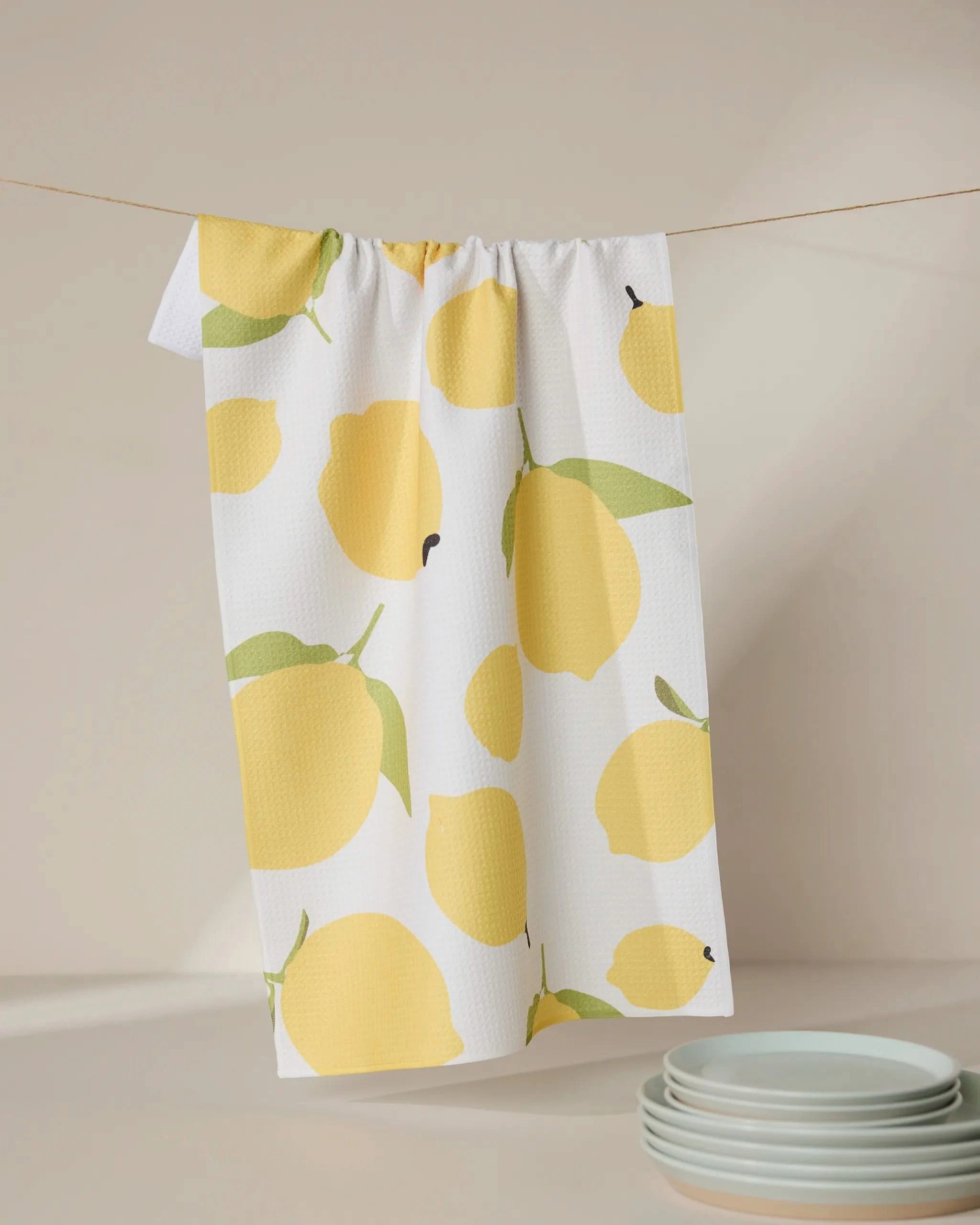 The GEOMETRY Sunny Lemons tea towel, crafted from recycled materials and featuring a yellow lemon print, hangs on a line. Light blue plates are stacked on a softly lit, neutral countertop in the corner.