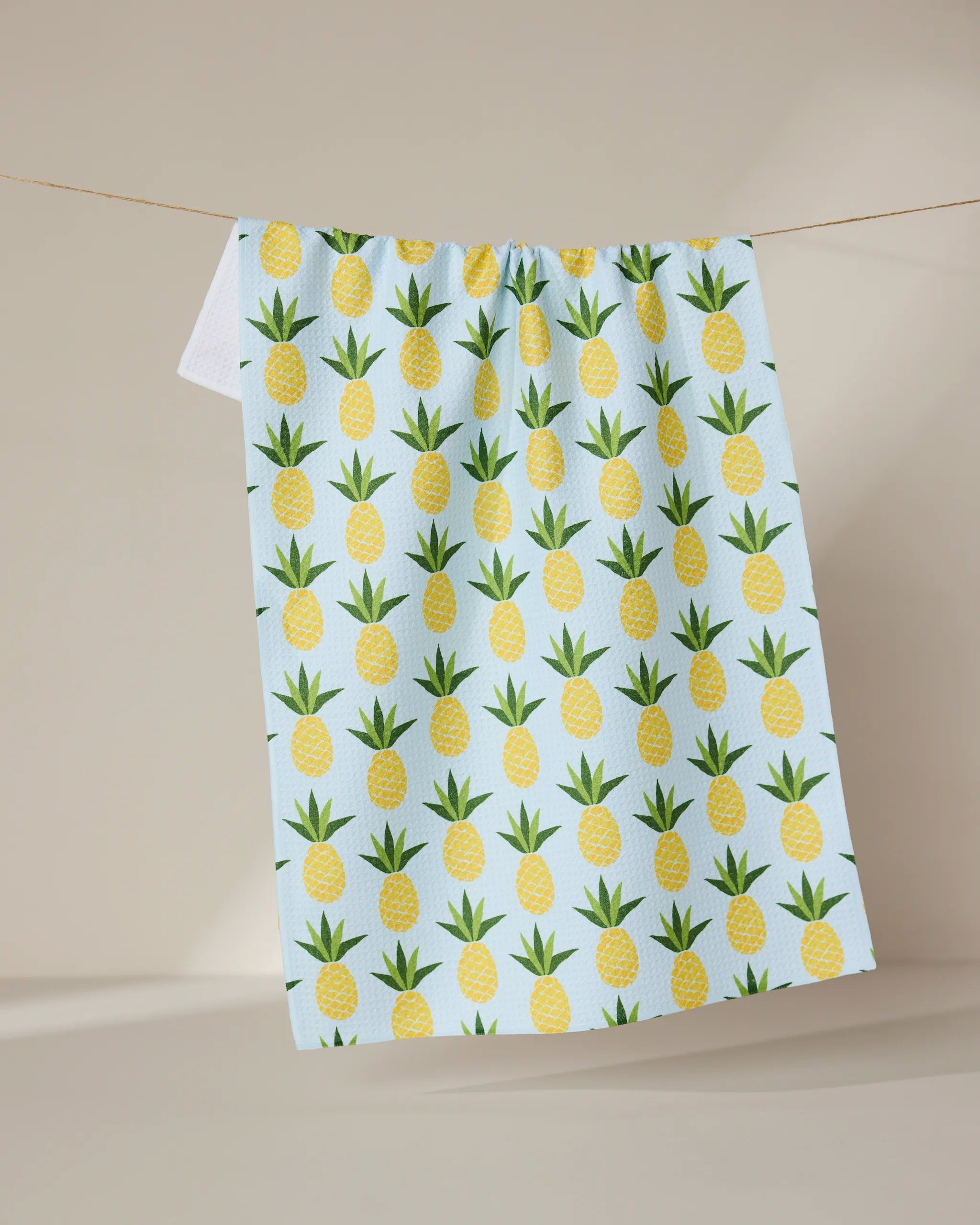 The Sweet Pineapple tea towel by GEOMETRY, featuring a light blue color with a repeating pattern of yellow pineapples and green leaves, hangs from a thin line against a neutral beige backdrop.