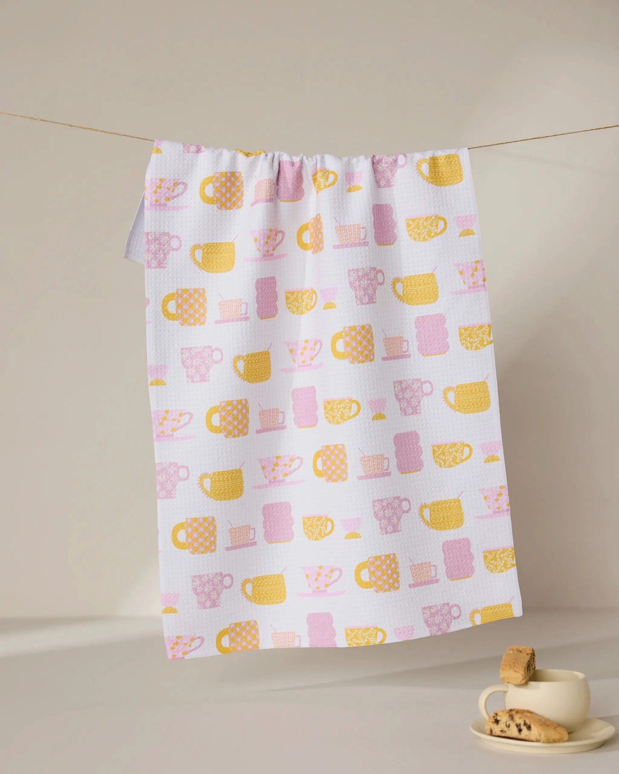The GEOMETRY Talk Over Tea towel, made from recycled materials and featuring yellow and pink mugs and cups, hangs on a line above a beige cup and saucer with cookies, set on a light surface against a neutral background.