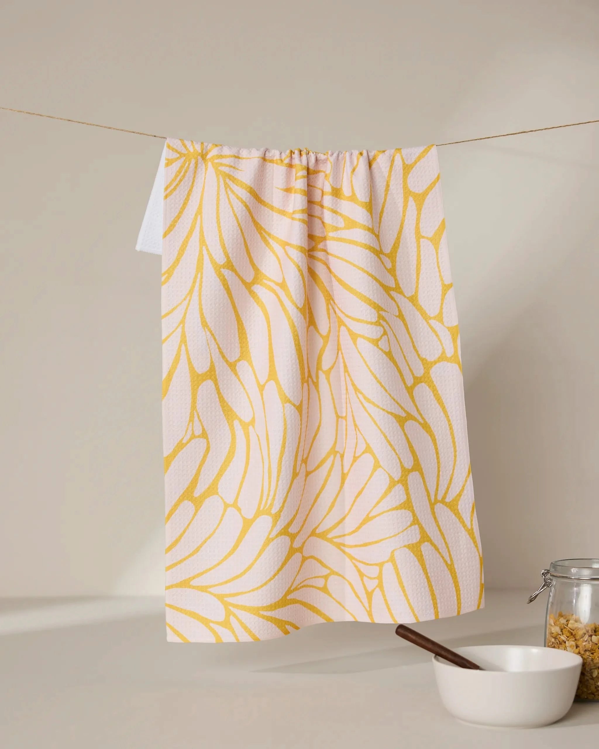 The GEOMETRY Tenderhearted tea towel, featuring a yellow abstract floral pattern, hangs on a string above a white bowl with a wooden spoon and a glass jar of cereal on a beige surface against a neutral background.
