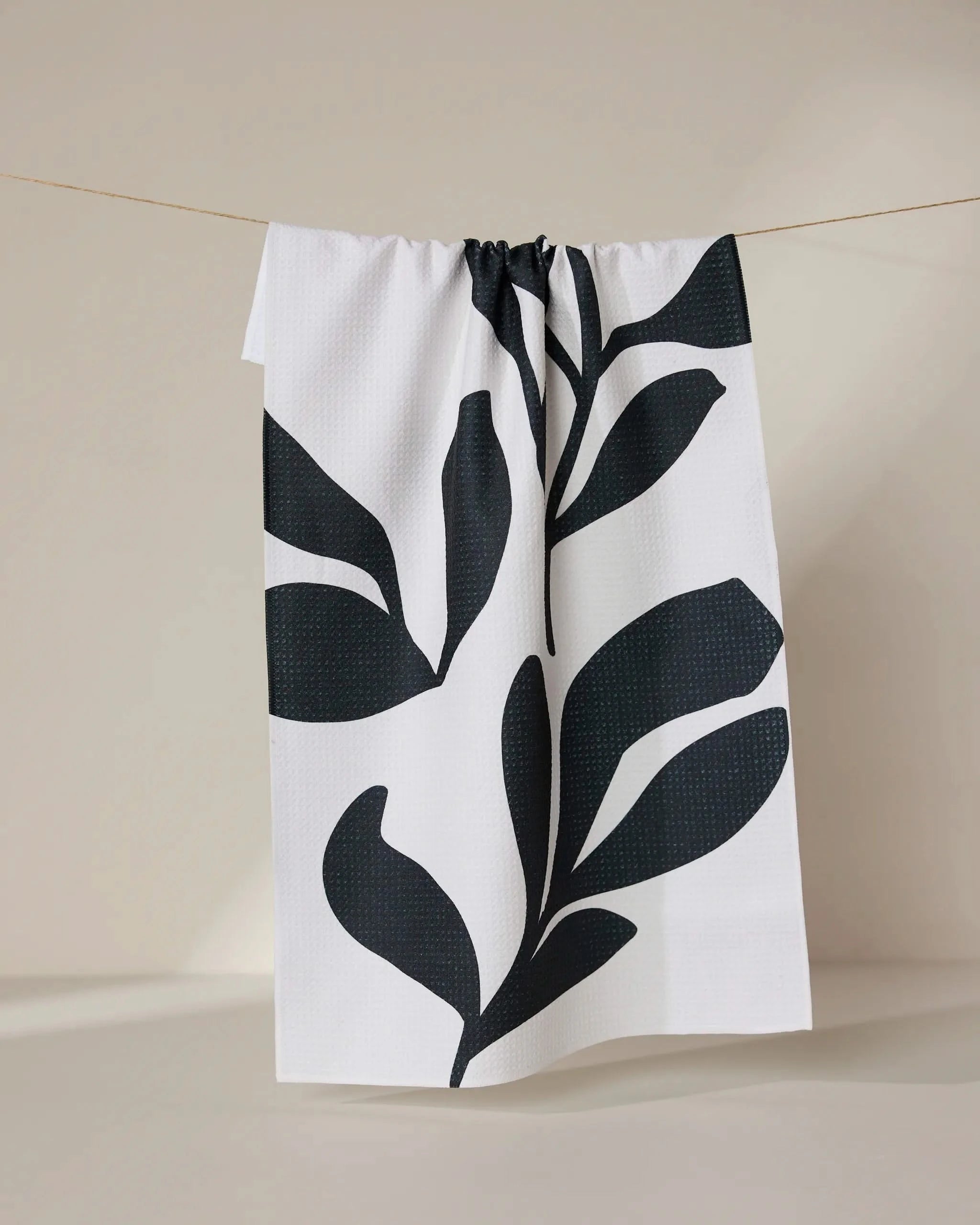 The GEOMETRY Threes tea towel, made from recycled materials, features large black abstract leaf patterns on white and hangs on a string against a plain, neutral background.