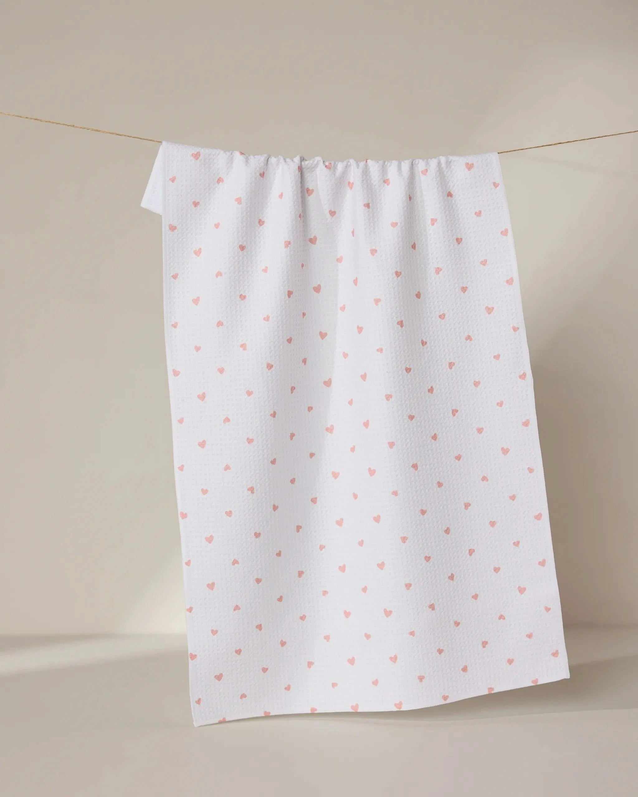 The GEOMETRY Tiny Hearts towel, ultra-absorbent and white with a pattern of small pink hearts, hangs on a clothesline against a neutral beige background.