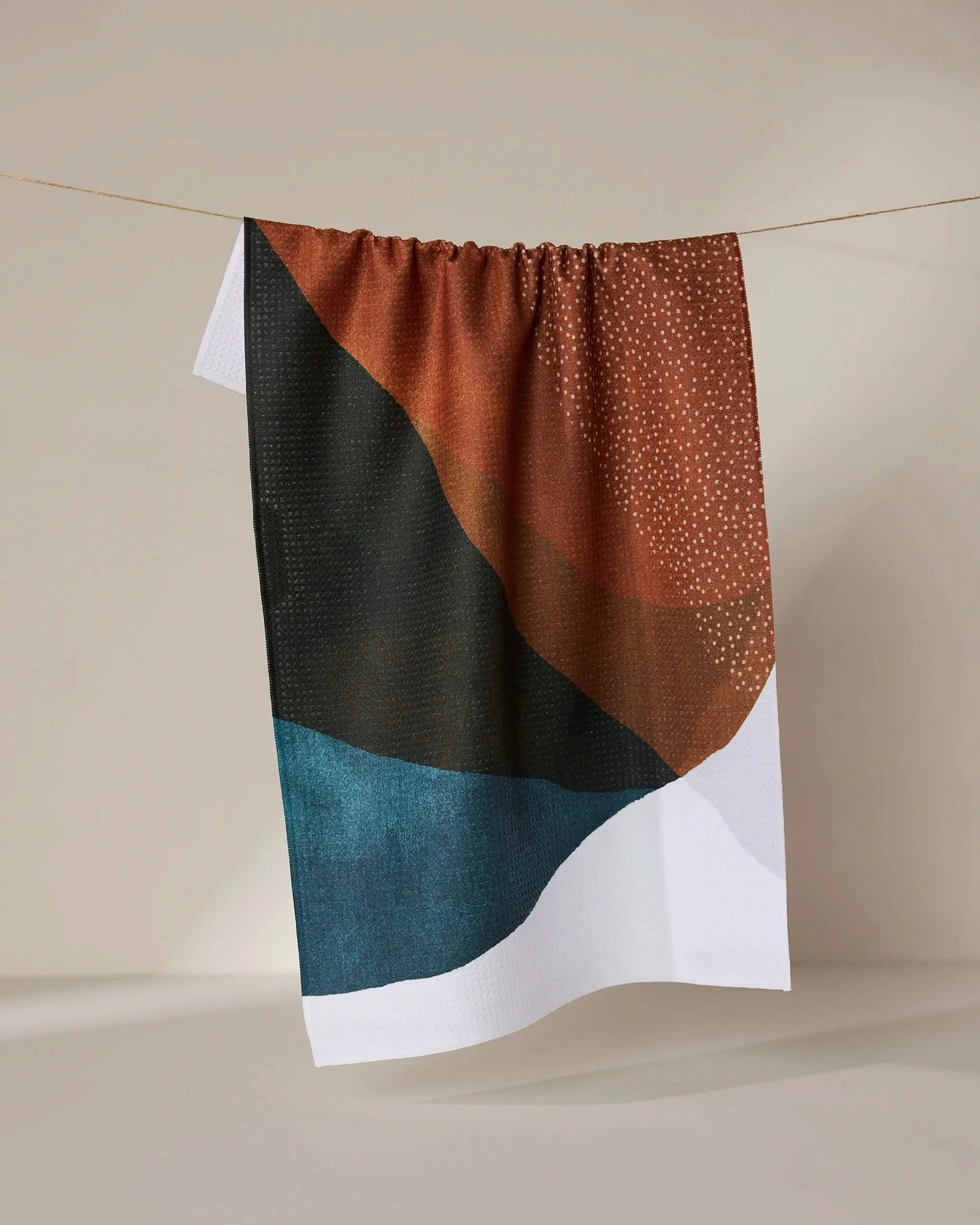 The GEOMETRY Tres recycled kitchen towel, with an abstract design in brown, black, teal, and white plus small white dots, hangs on a line against a neutral background.