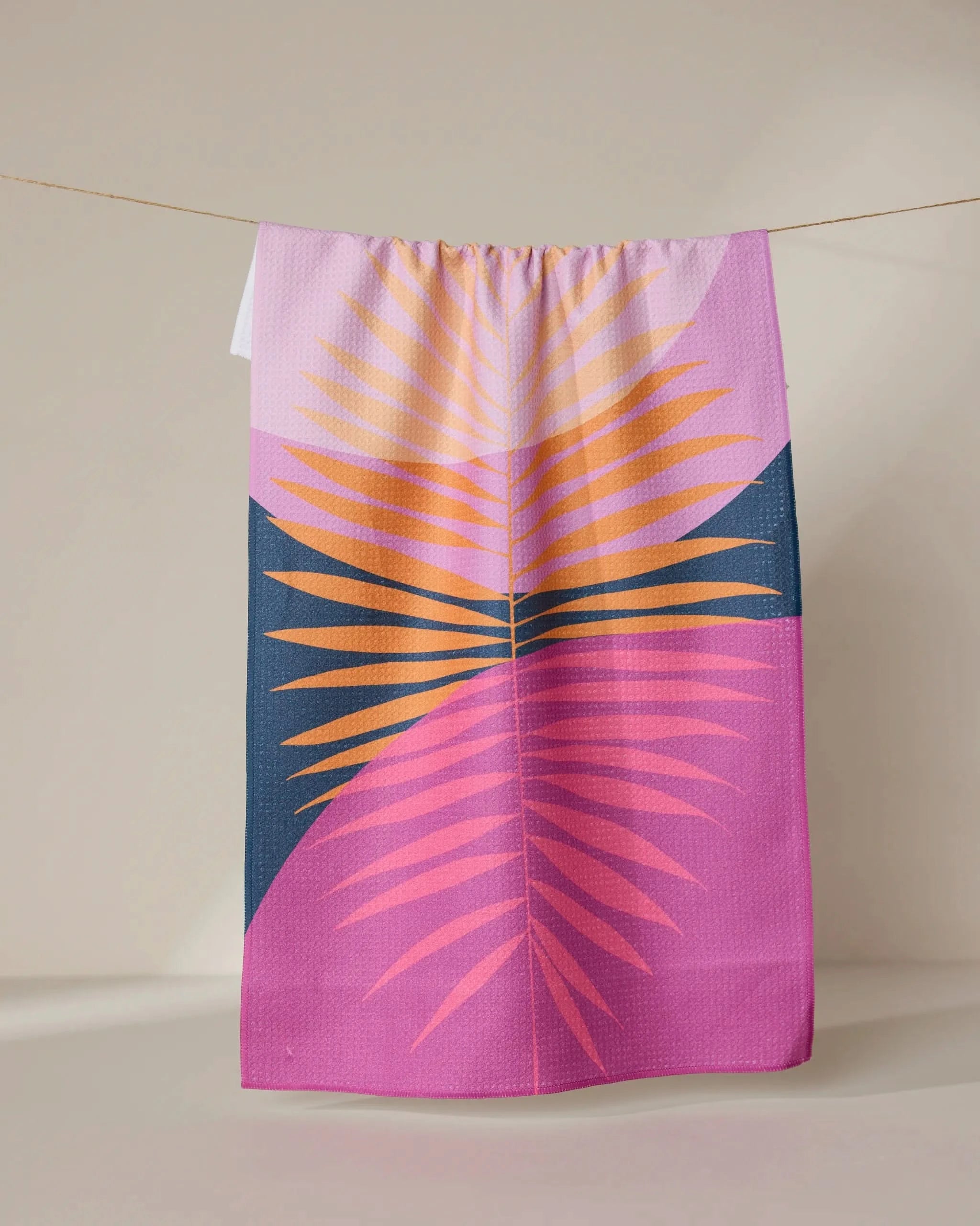 The GEOMETRY Tropical Summer tea towel, made with Geoweave fabric and featuring vibrant palm leaves on a purple and dark blue background, hangs on a clothesline against a neutral wall.