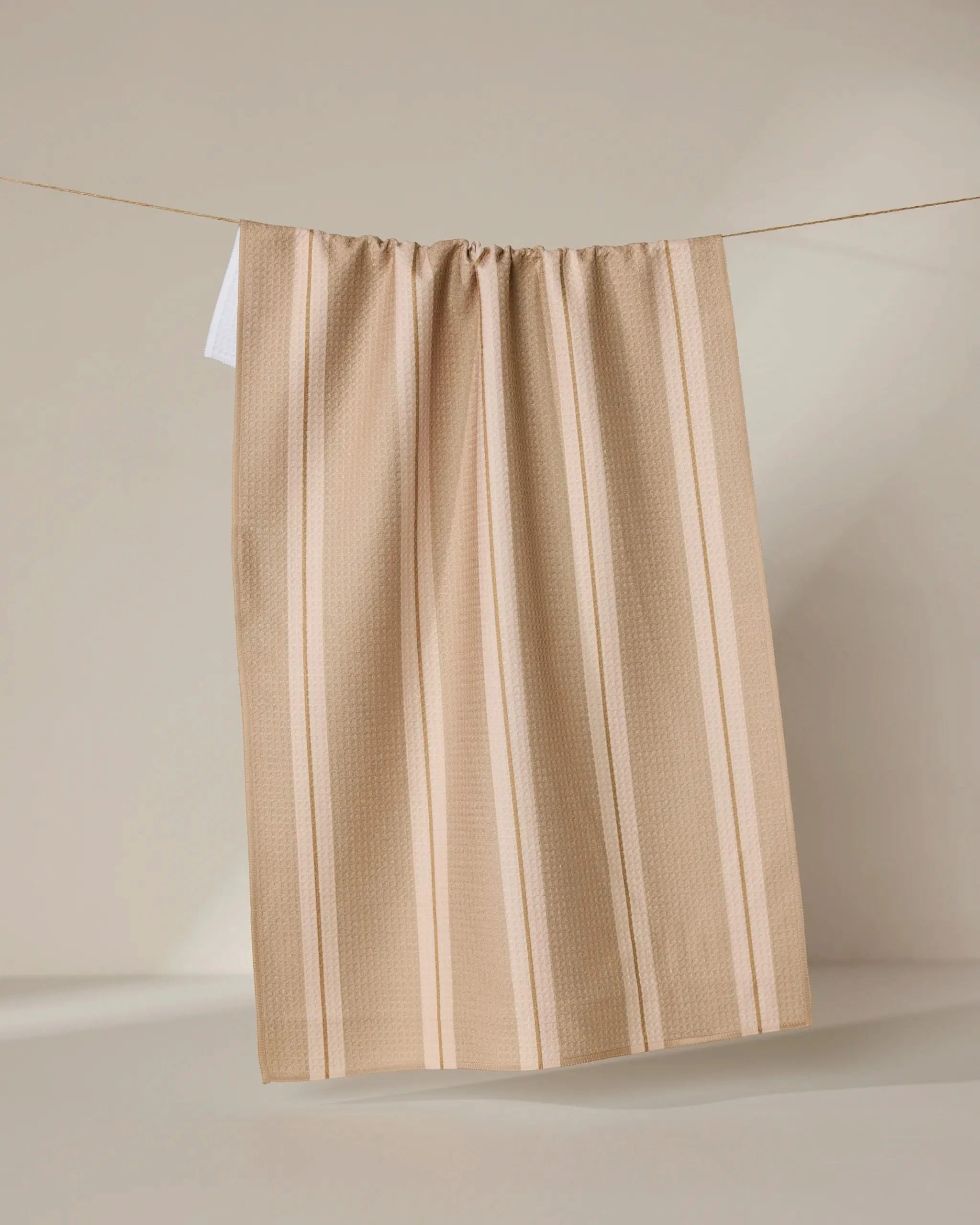 The Vanilla Chai tea towel by GEOMETRY, featuring beige and cream stripes and made from recycled materials, hangs on a clothesline against a neutral background with soft light casting gentle shadows.