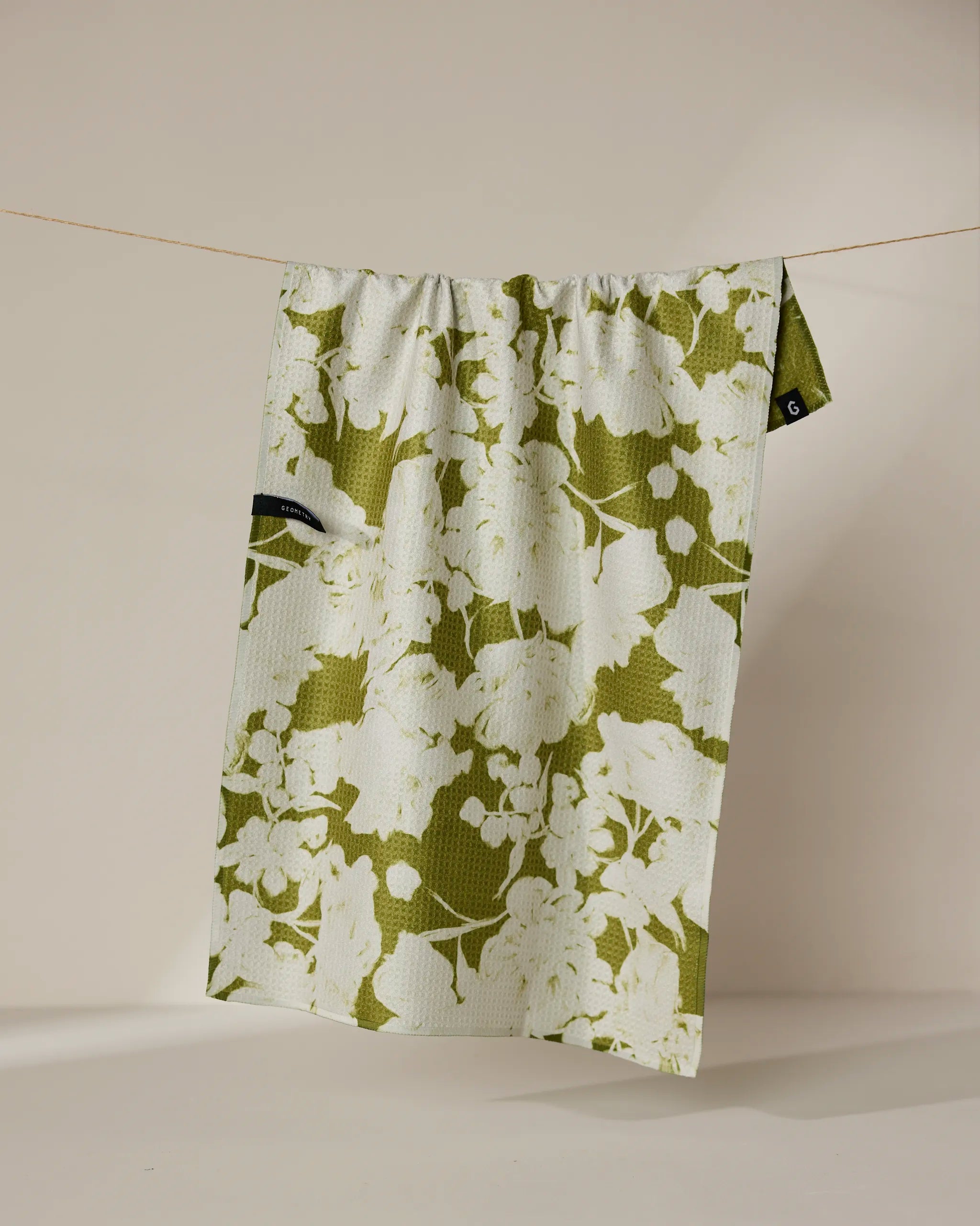 The Geometry “Sweet Green” recycled kitchen towel, featuring a green and white floral pattern and a small black tag, hangs on a thin string against a plain beige background.