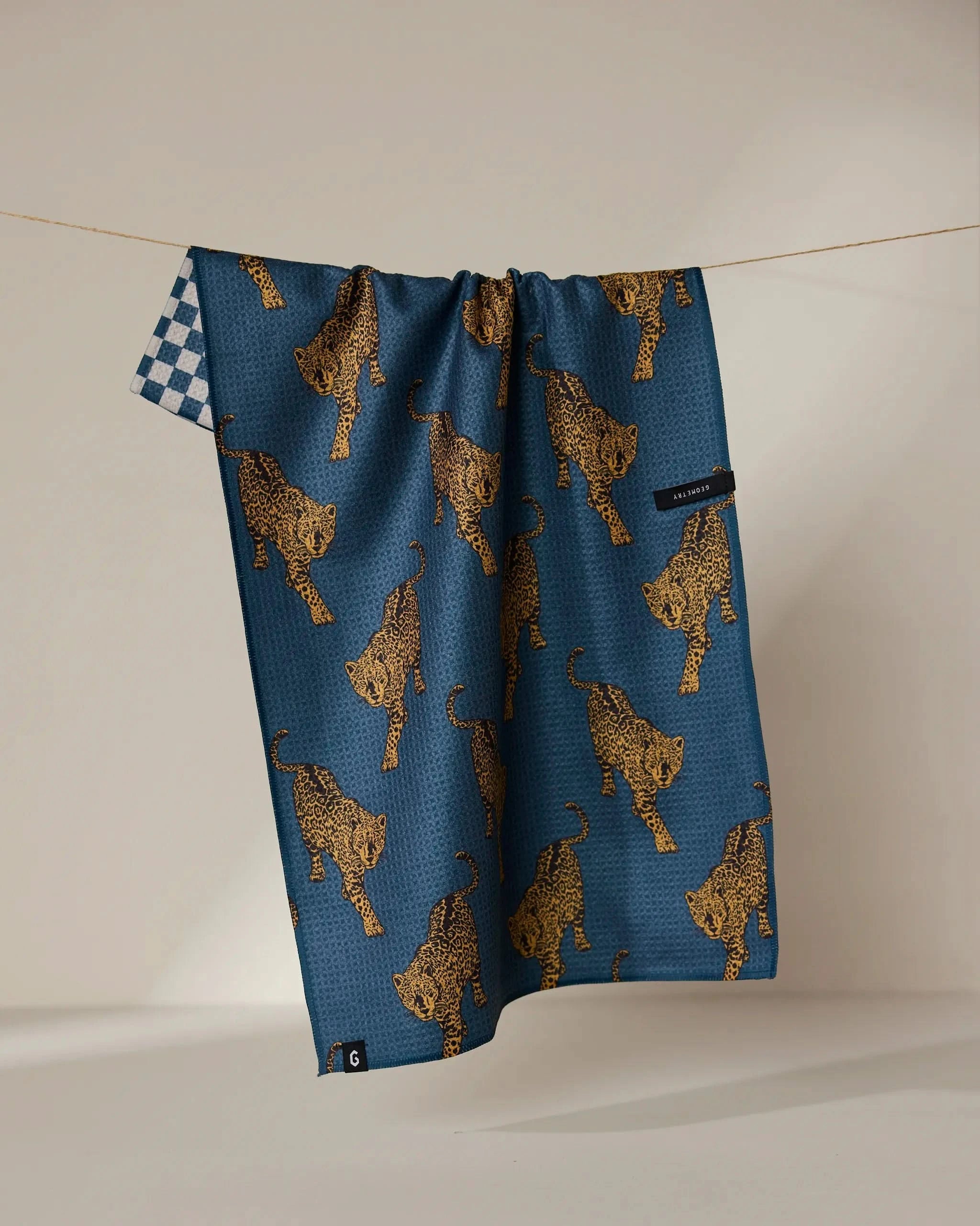 The Geometry Leopard Lovin tea towel, featuring blue with yellow leopards on one side and a blue and white checkerboard on the other, hangs on a clothesline—adding vibrant style and quick-drying practicality to your kitchen.
