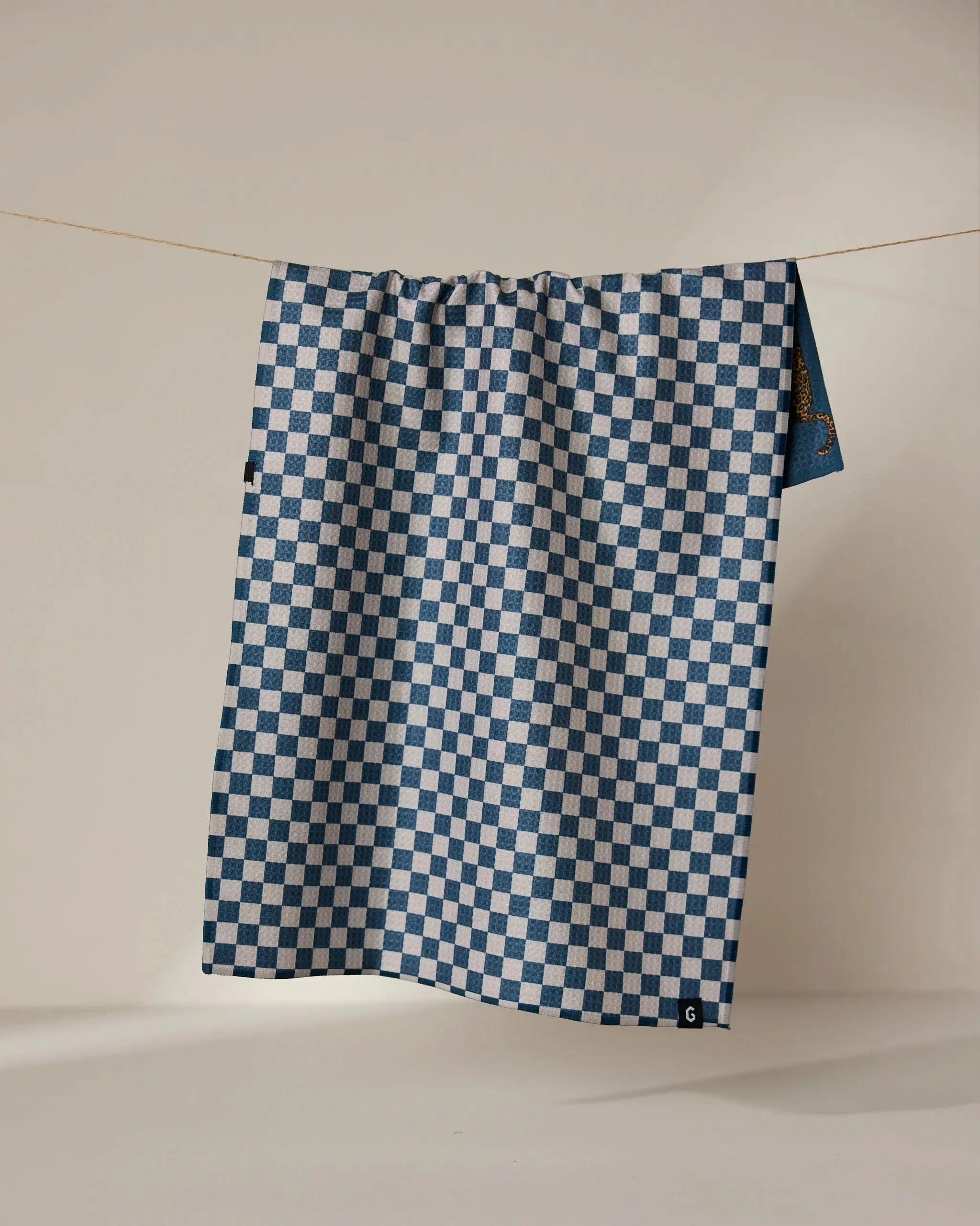 A reusable Leopard Lovin kitchen towel by Geometry hangs over a thin rope against a light background. The double-sided tea towel features a black tag with a white letter C on its corner.