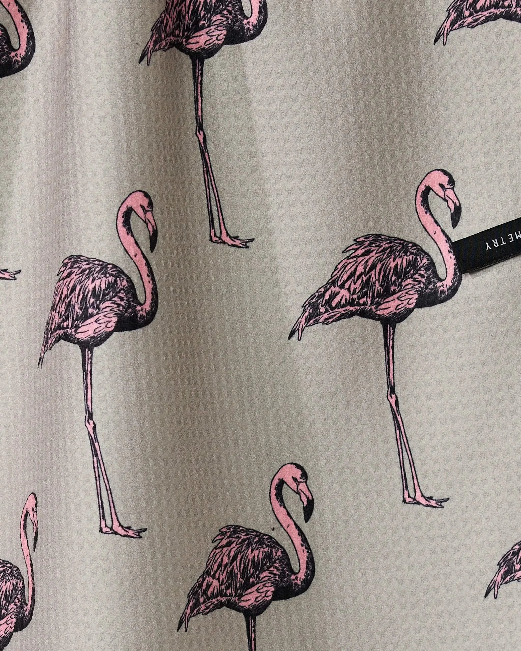 The Geometry Flamingo Dance tea towel features gray textured fabric with illustrated pink flamingos in various poses and a small black tag on the right side.