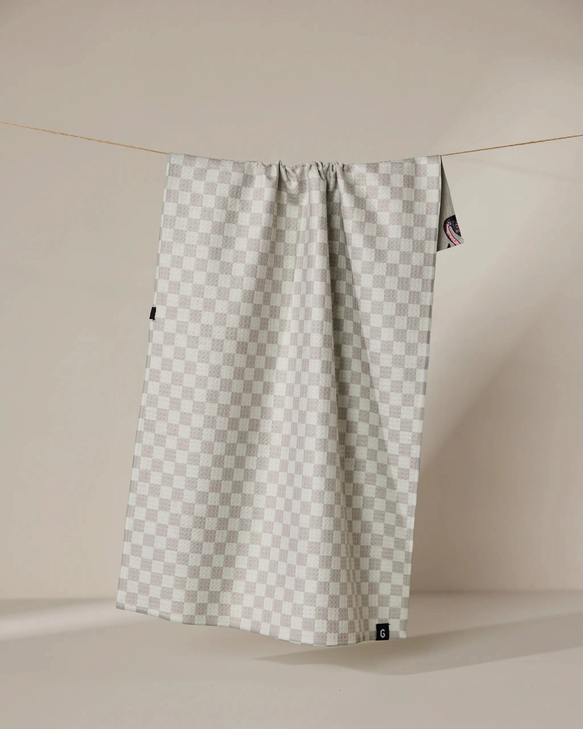 The Geometry Flamingo Dance double-sided tea towel, featuring a light gray and white checkered pattern, hangs on a thin clothesline against a beige background with subtle shadows.