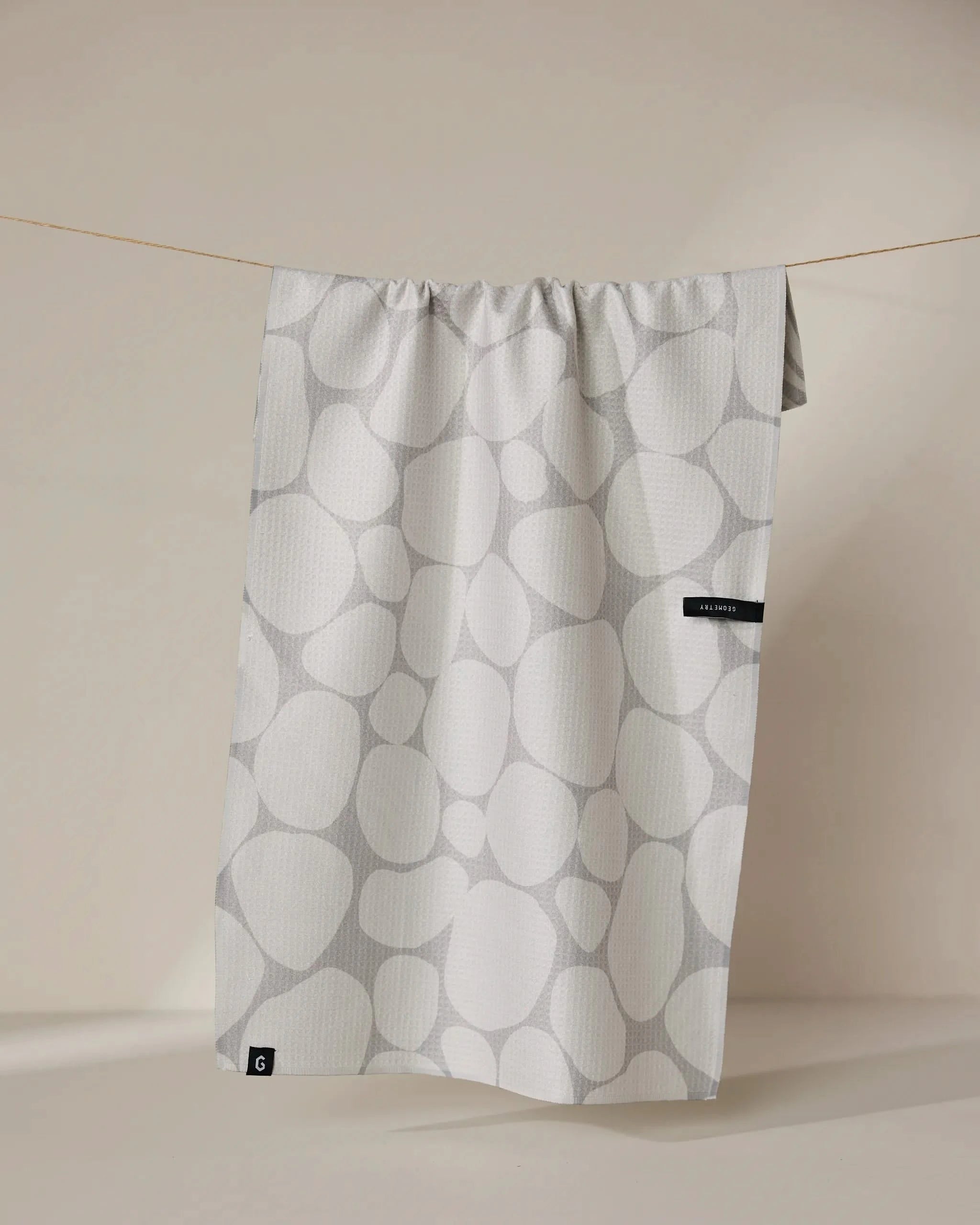The Geometry Stone recycled kitchen towel, featuring a light gray pebble pattern and two black tags on one side, hangs from a thin string against a beige background.