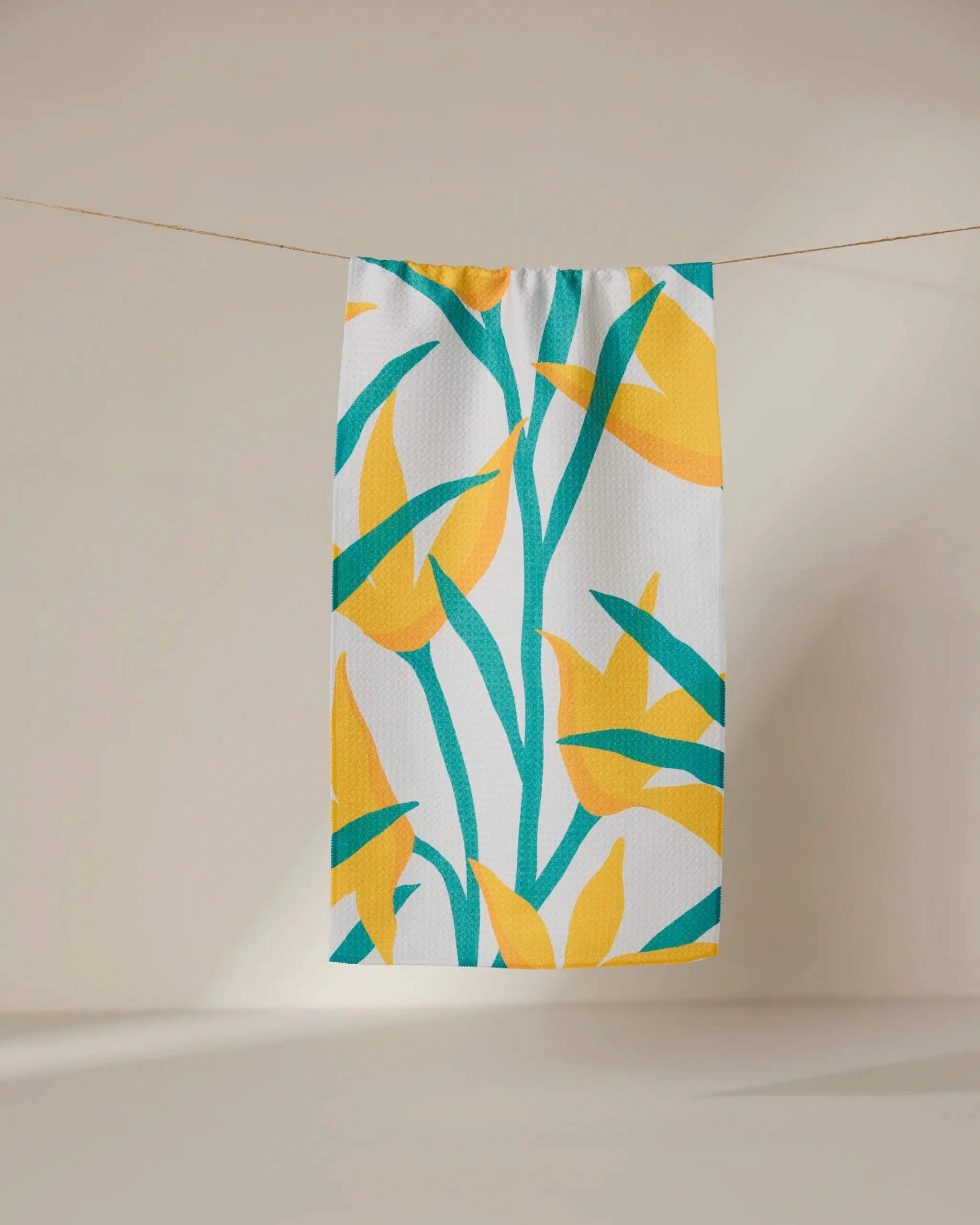 The GEOMETRY Growth towel, ultra-absorbent and white with vibrant yellow and green tropical leaf print, hangs on a clothesline against a neutral background.