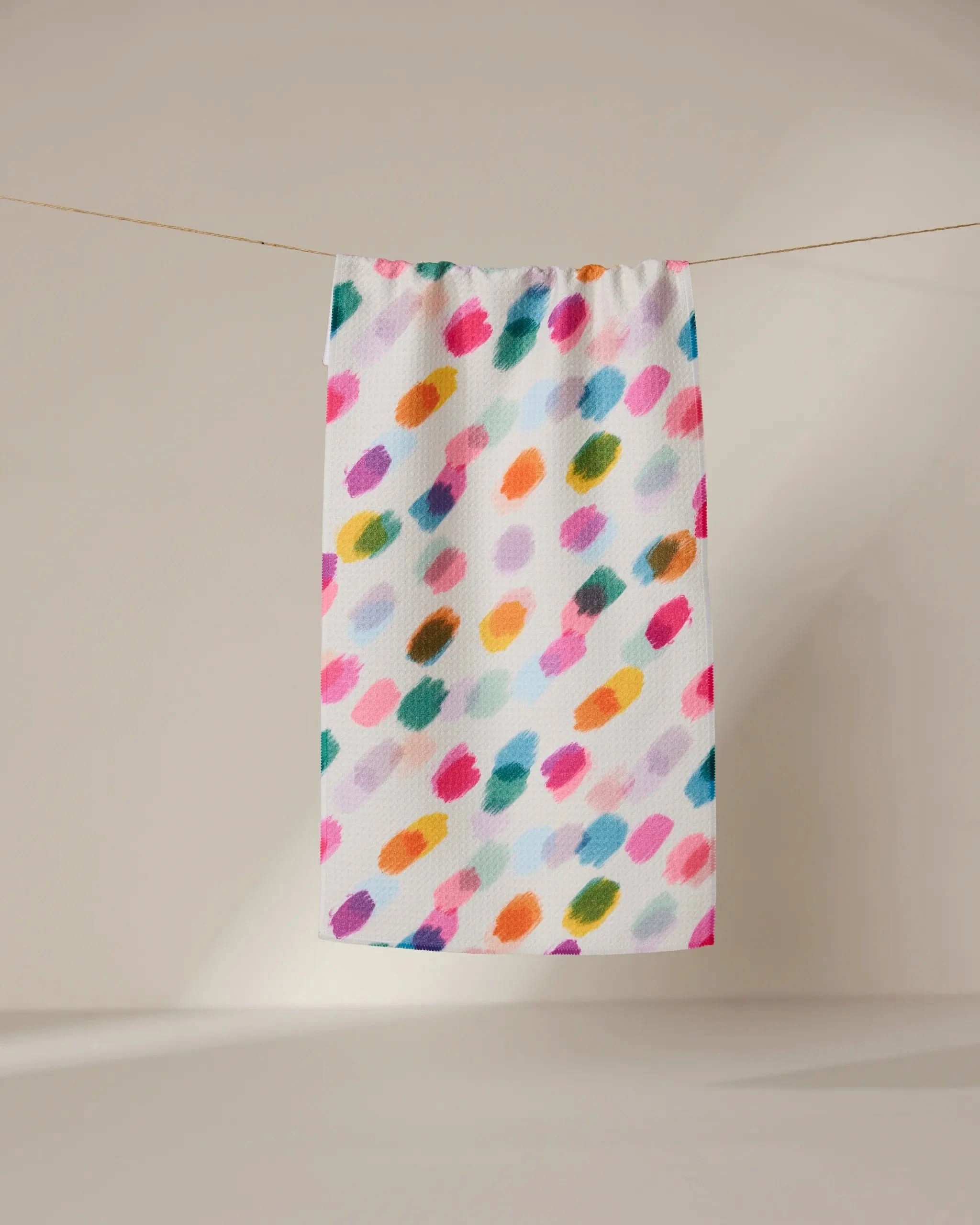 The GEOMETRY Downpour towel, ultra-absorbent and white with vibrant pink, orange, green, blue, and purple brushstroke patterns, hangs on a string against a softly lit plain background.