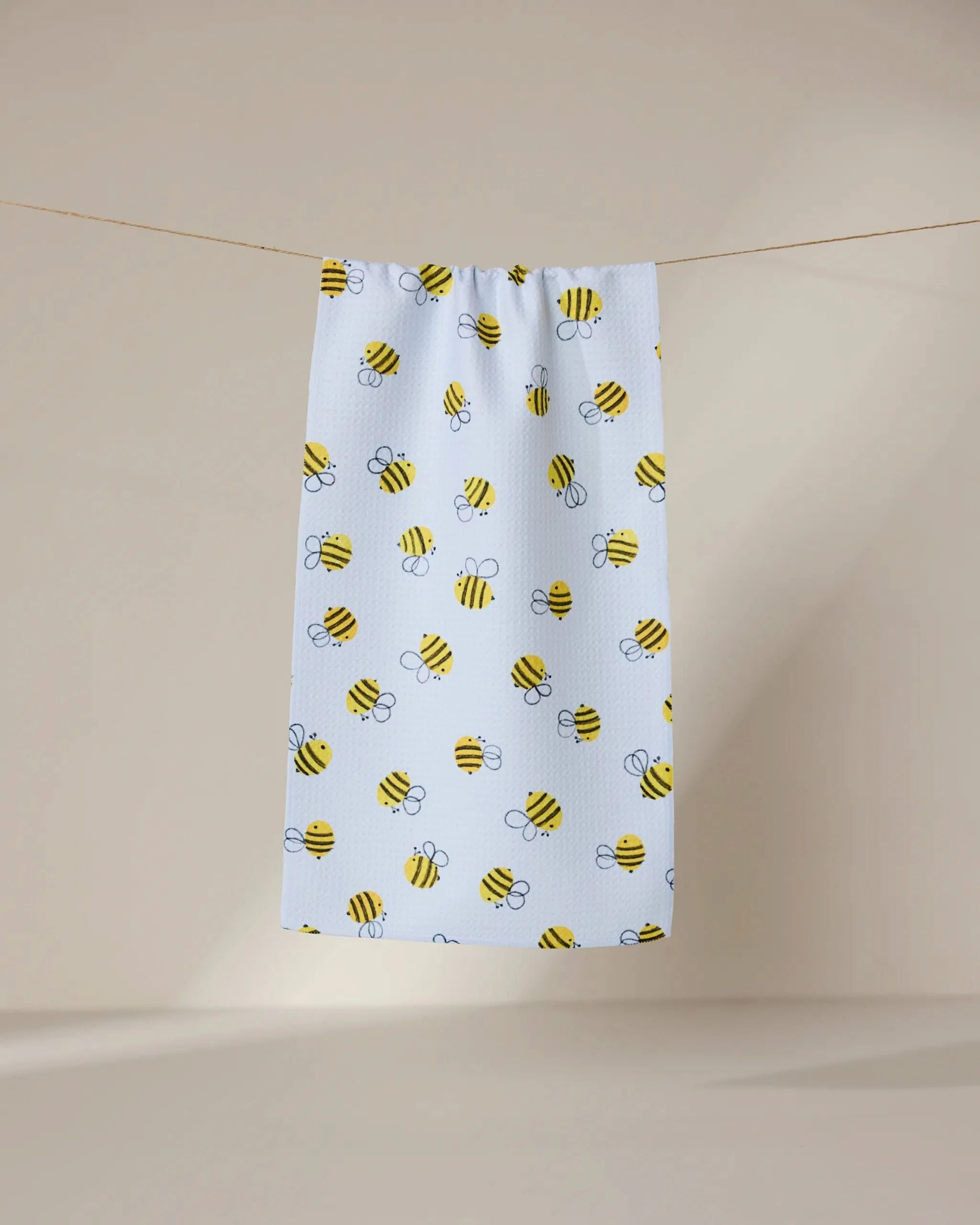 The GEOMETRY Buzzing towel, ultra-absorbent and white with yellow and black cartoon bee patterns, hangs on a clothesline against a plain beige background.