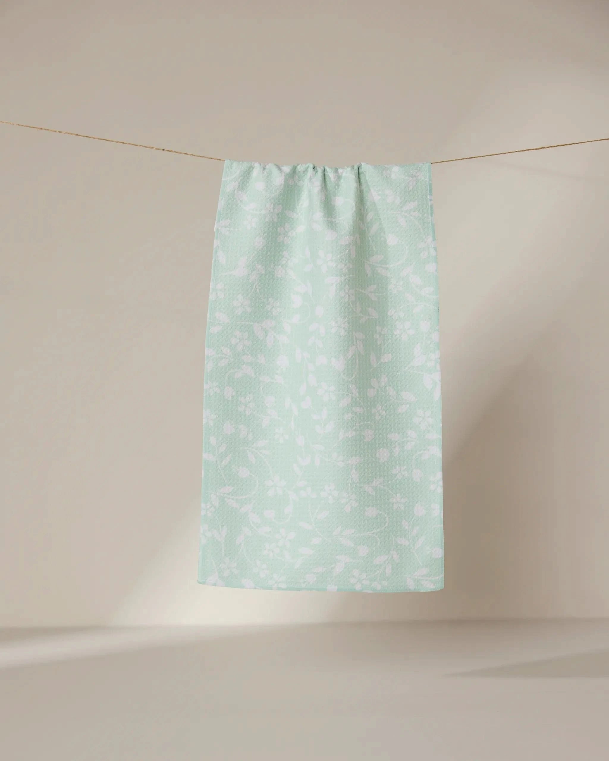 The GEOMETRY Dandy towel, in light green with a subtle white floral pattern, hangs on a clothesline against a neutral beige background.