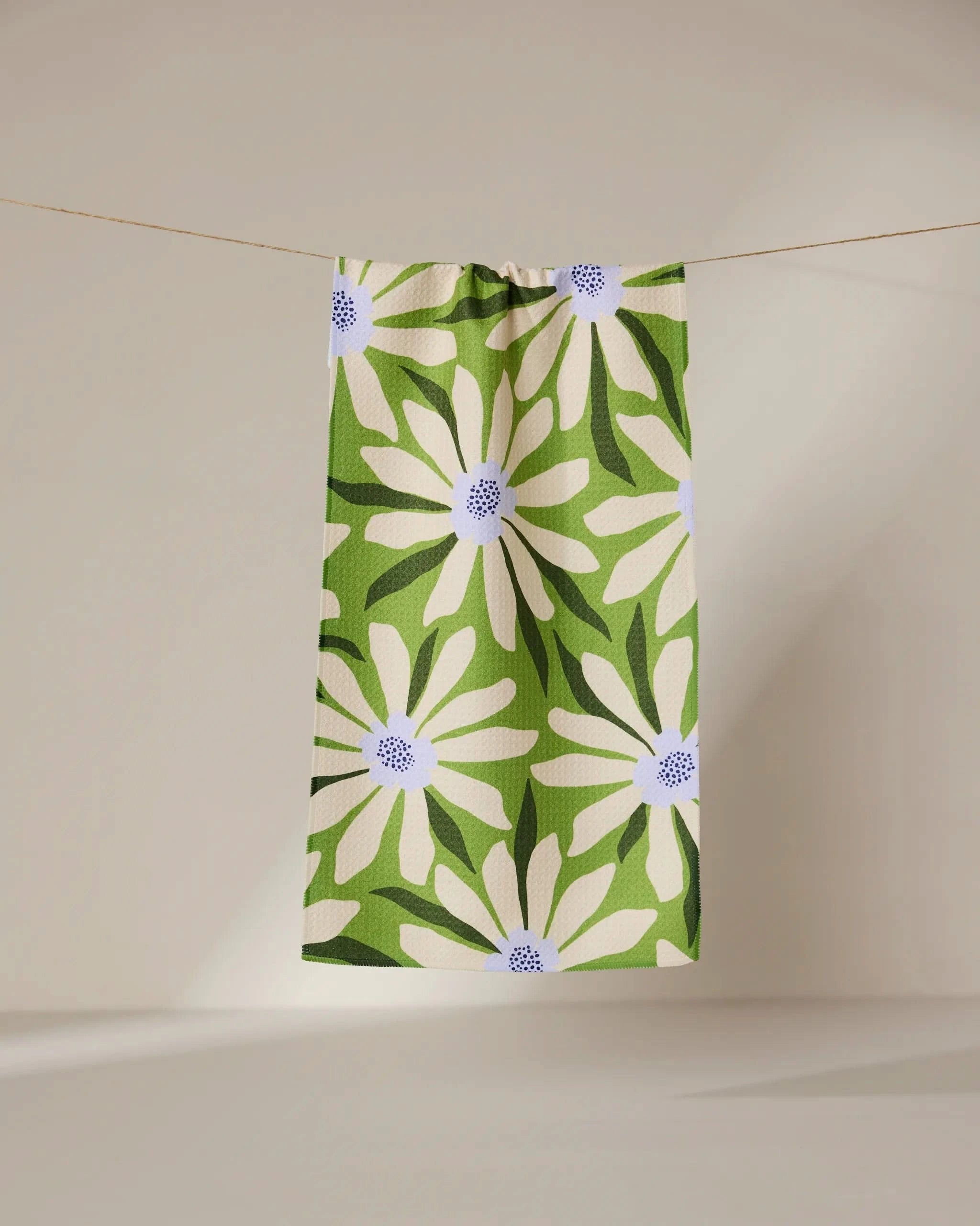 The GEOMETRY Bliss and Bloom green recycled kitchen towel, featuring a striking white daisy with a blue center, hangs on a clothesline against a neutral background.
