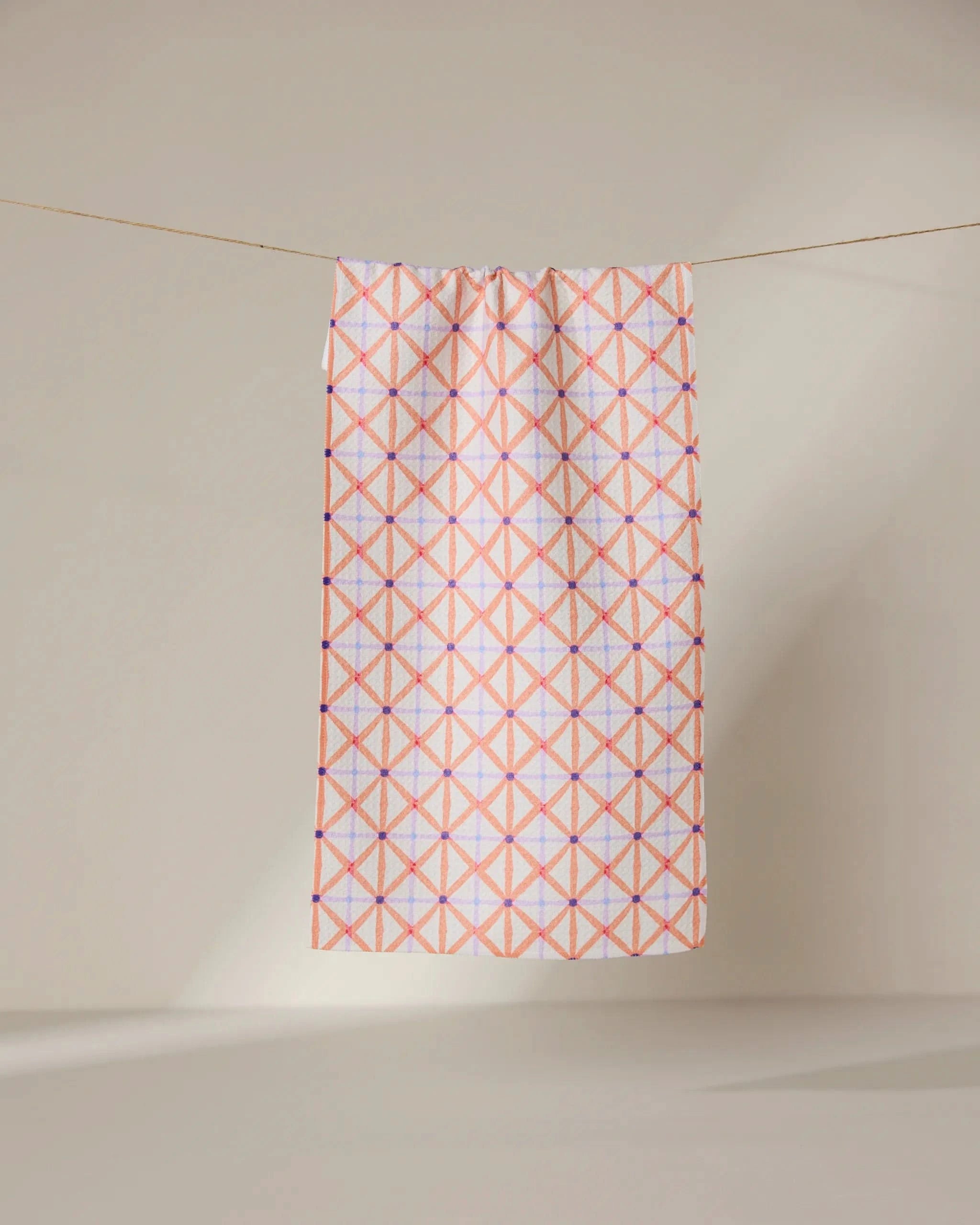 The GEOMETRY Anew towel features ultra-absorbent fabric and a geometric pattern of intersecting pink and orange lines forming diamonds, shown hanging on a line against a neutral, light background.