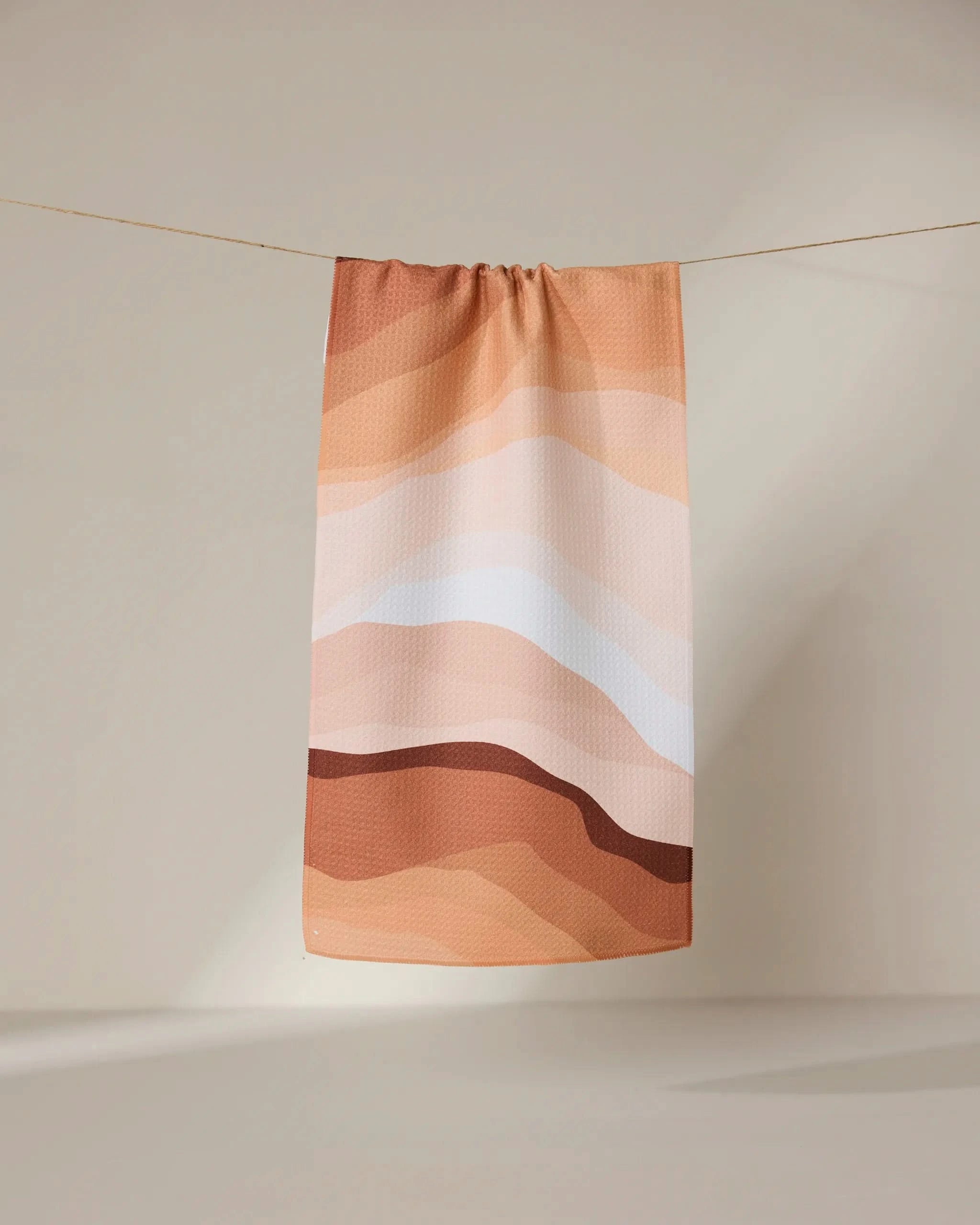 The GEOMETRY Levels of Fall towel, featuring ultra-absorbent fabric and abstract wavy stripes in brown, beige, tan, and white, hangs on a clothesline against a neutral background.