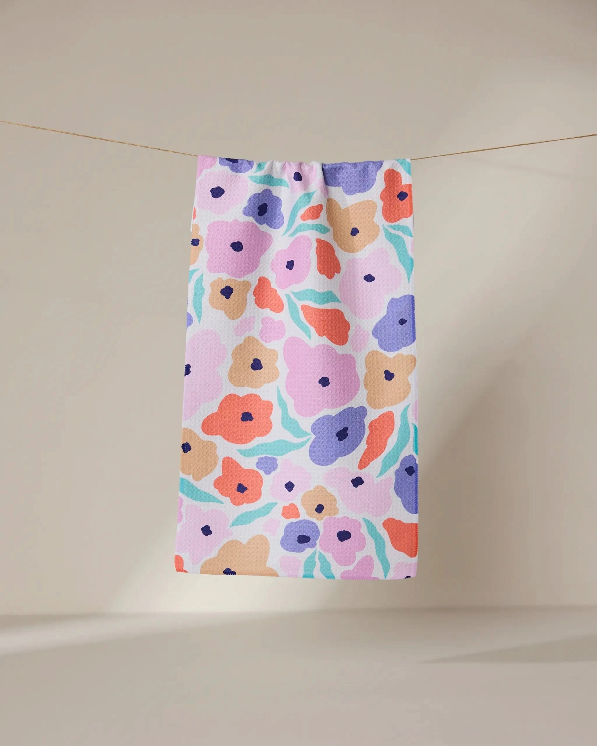 The Blossoming Love towel by GEOMETRY, featuring a bold abstract floral design in purple, orange, pink, and blue hues, hangs from a line against a neutral backdrop.