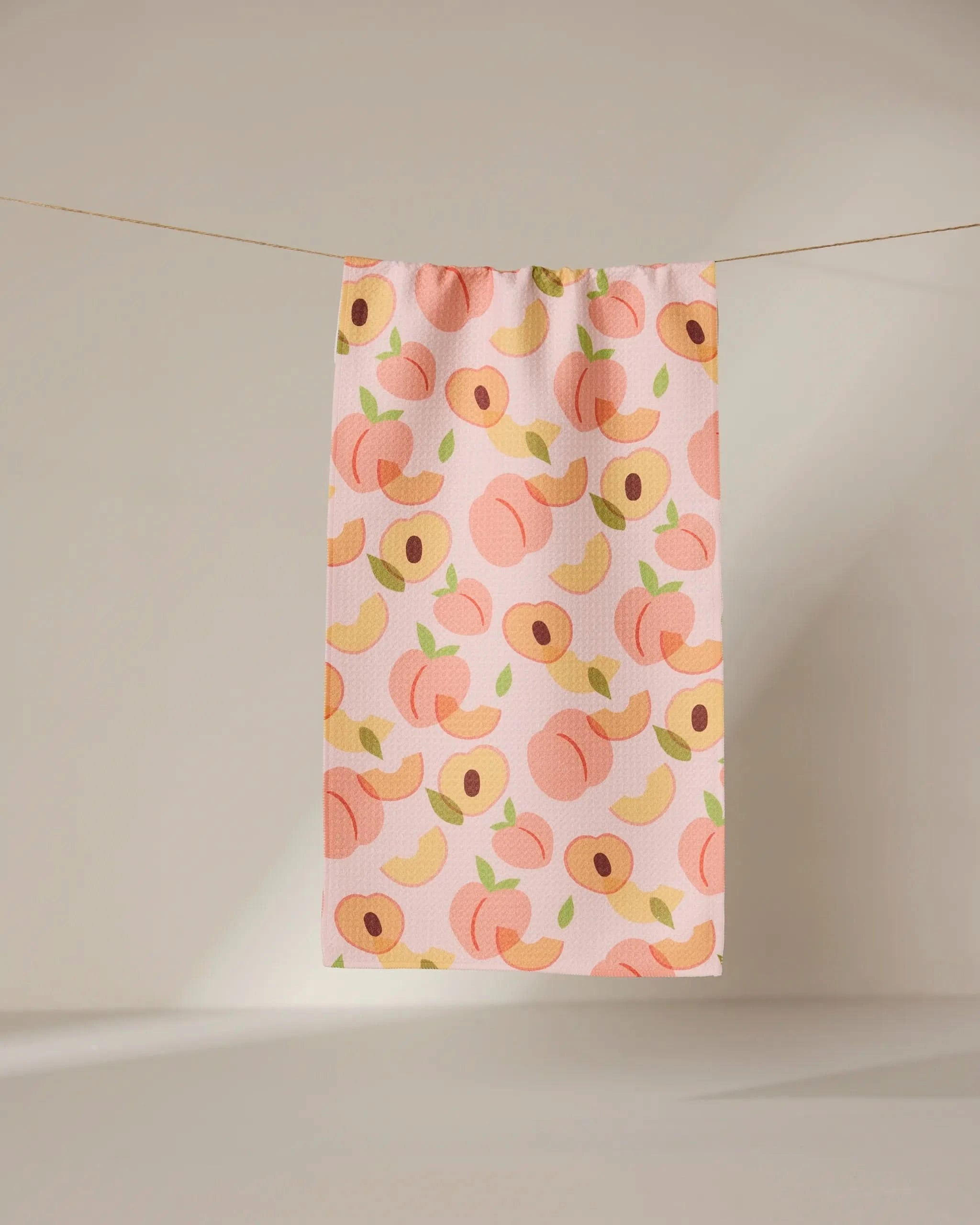 The GEOMETRY Peaches towel, in light pink and adorned with whole and sliced peaches in pink, yellow, and green, hangs on a clothesline against a neutral backdrop.