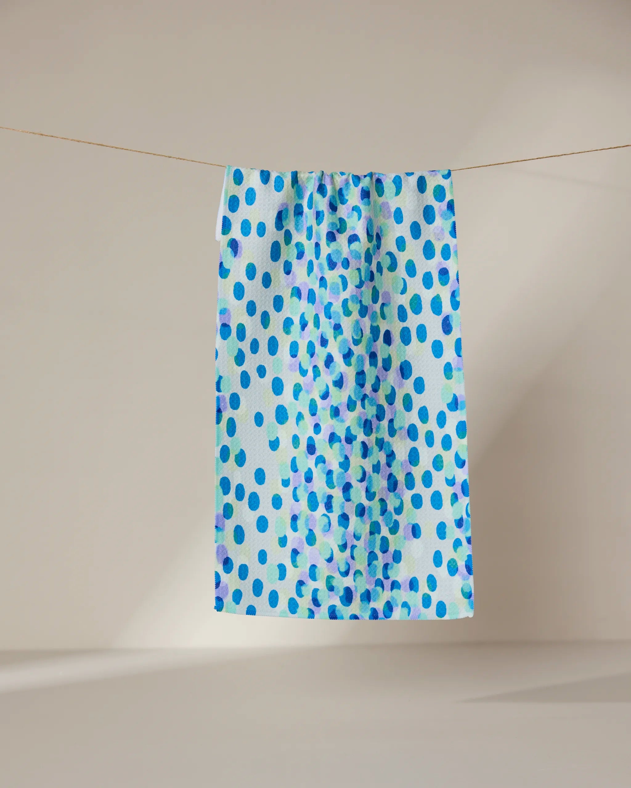The GEOMETRY Spotty Summer towel, featuring a blue polka dot design and ultra-absorbent rectangular shape, hangs on a clothesline against a softly lit neutral background.