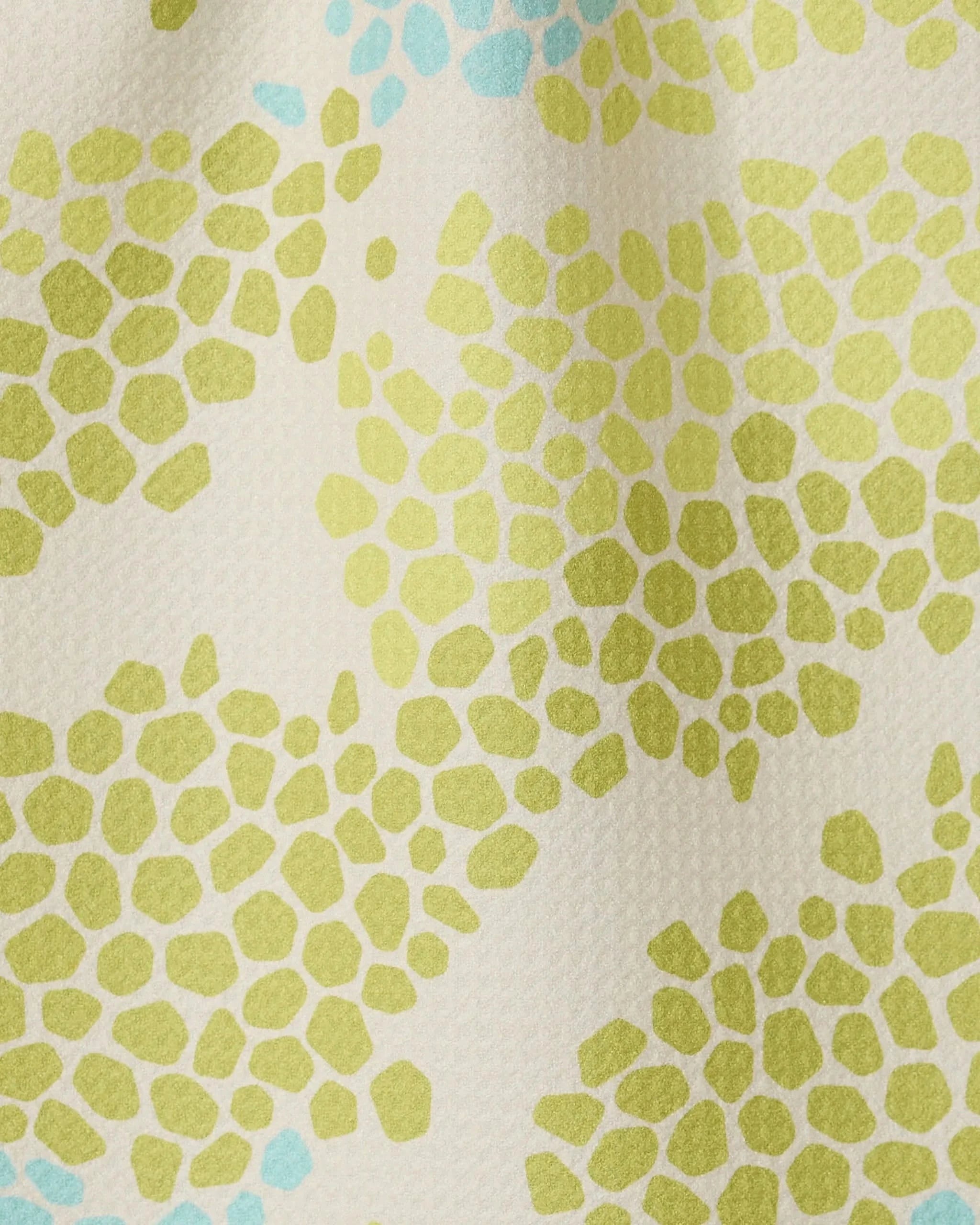 A close-up of GEOMETRY’s Geo Shell fabric shows clustered green and light blue irregular dots on a cream background, crafted from recycled materials and resembling abstract floral or pebble shapes.