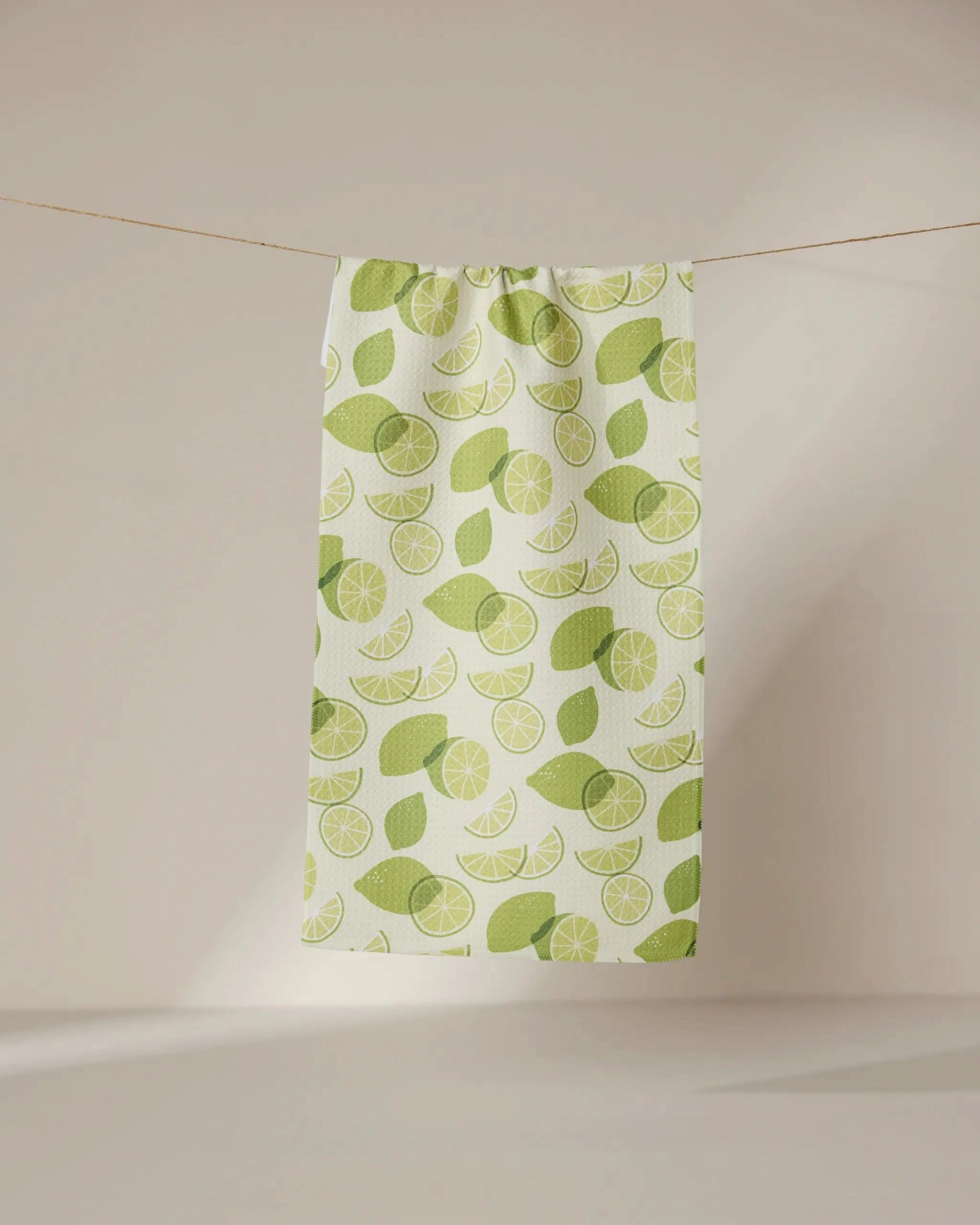 A white ultra-absorbent GEOMETRY towel named Limes, featuring a green lime and leaf pattern, hangs from a clothesline against a softly lit, plain background.