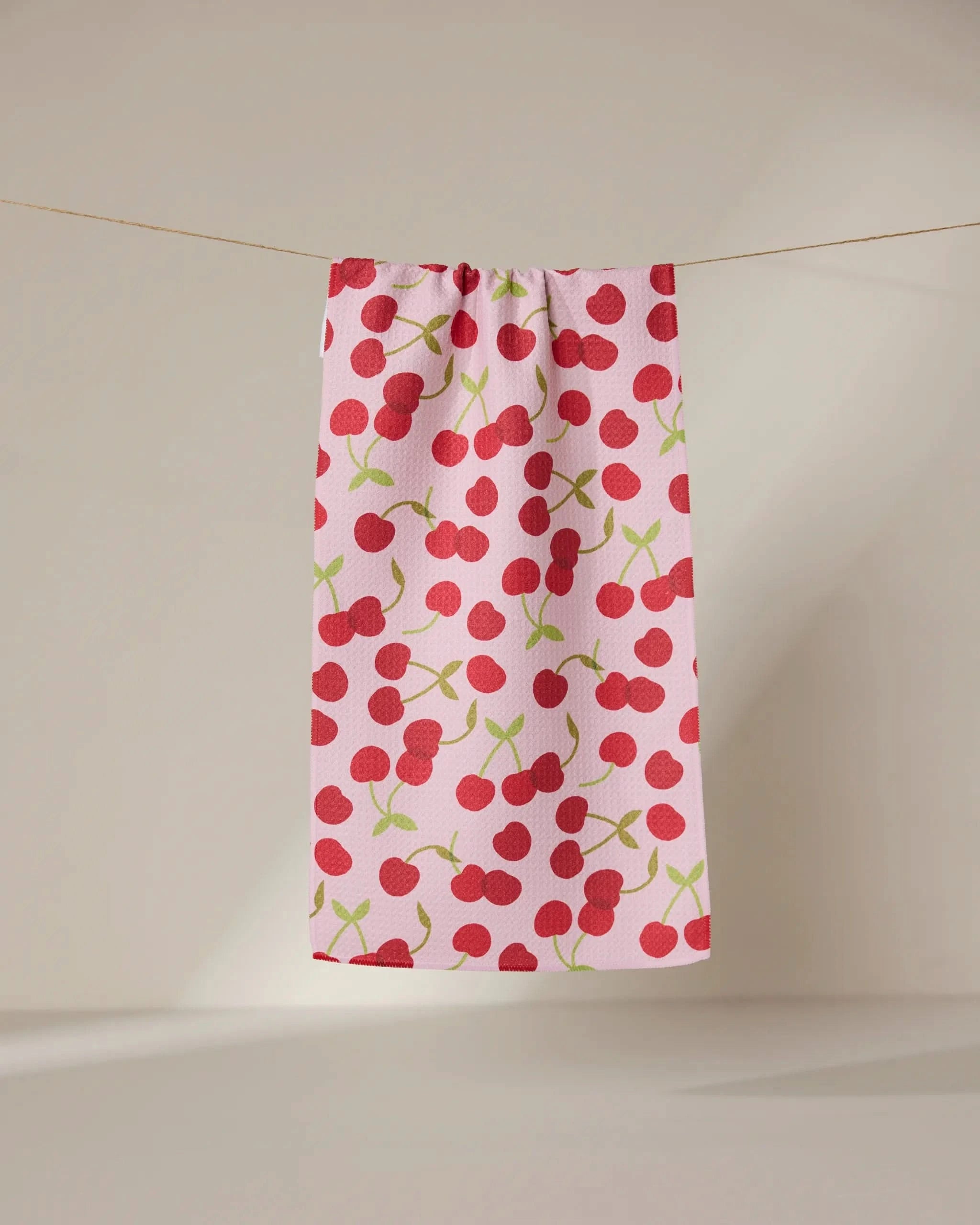 The GEOMETRY Cheery Cherries towel, ultra-absorbent and pink with red cherry and green stem patterns, hangs on a clothesline against a neutral background.