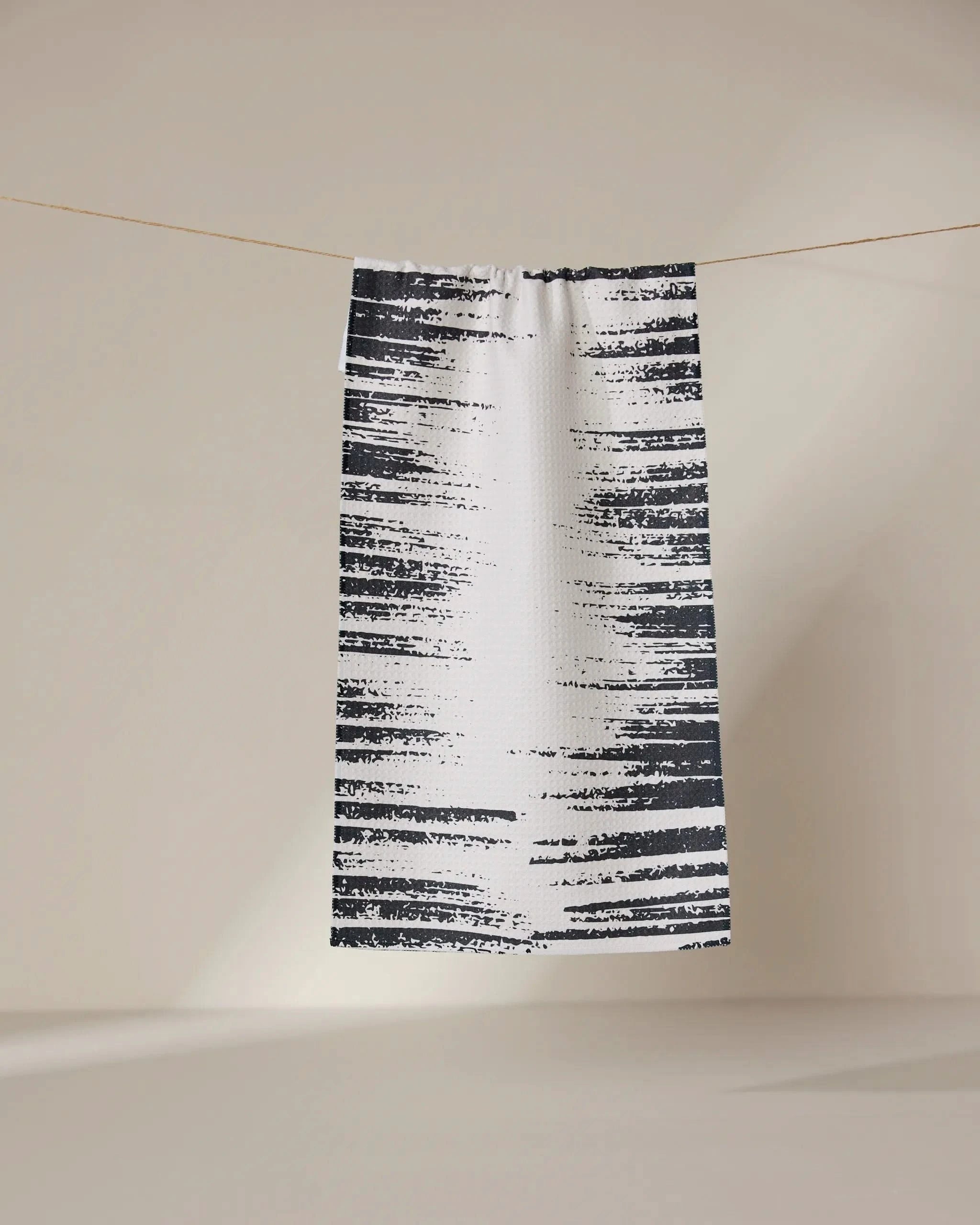 The GEOMETRY Worn Edges towel, featuring irregular black stripes on white, hangs on a clothesline in front of a neutral beige background.