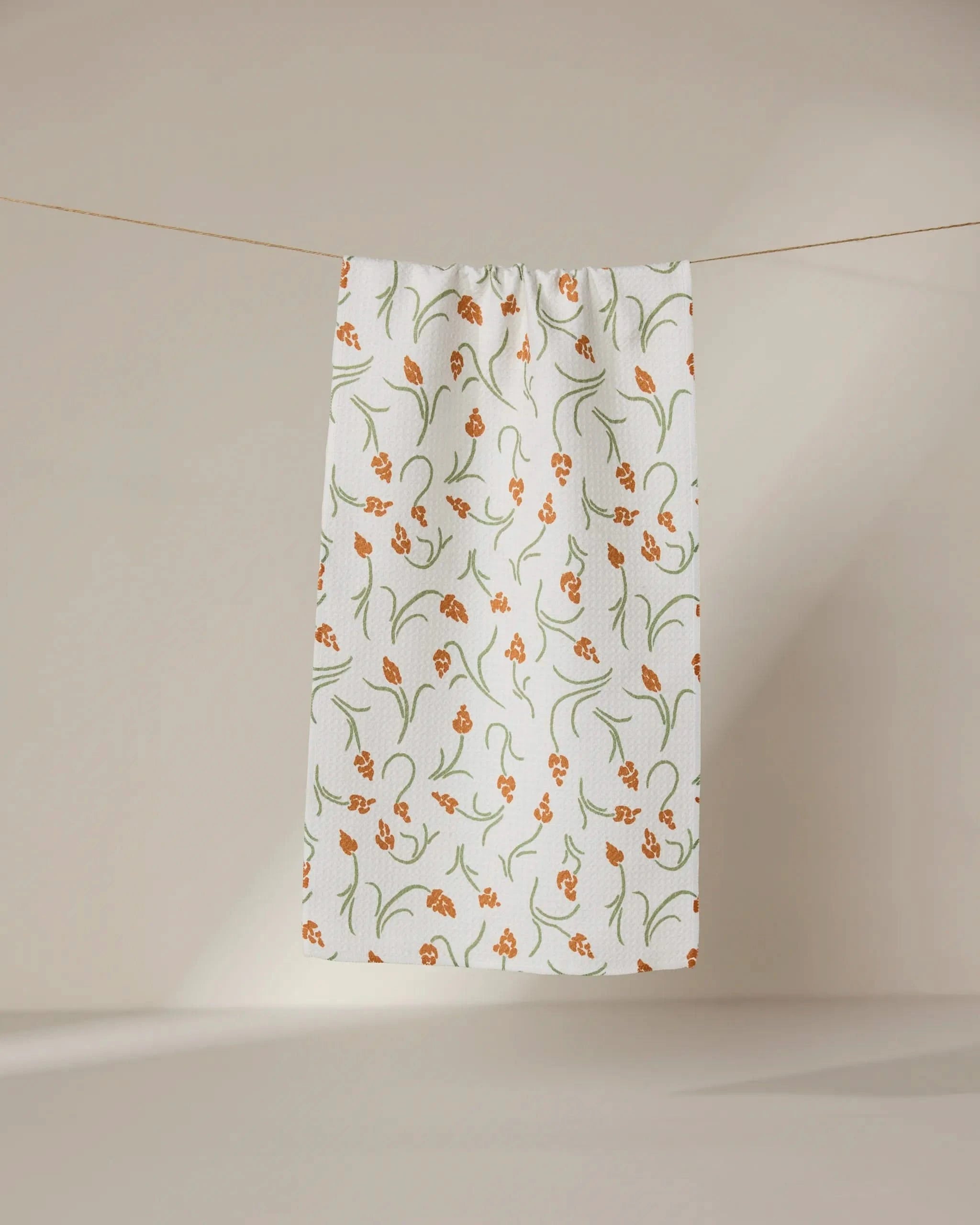 The GEOMETRY Crayon Floral ultra-absorbent white towel, featuring an orange and green floral design, hangs against a neutral beige background.