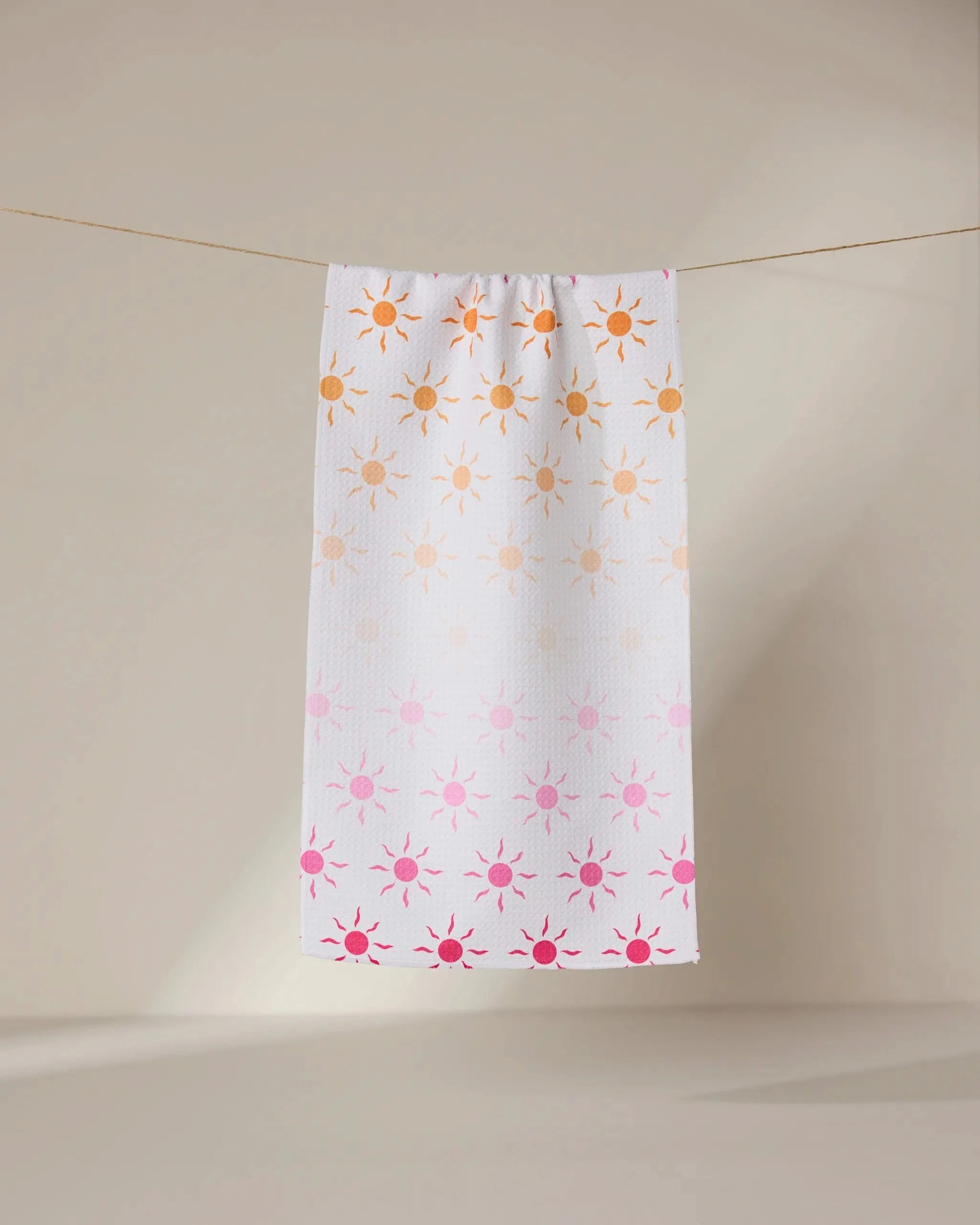 The GEOMETRY Fun in the Sun bar towel, featuring orange and pink sun patterns on white, hangs on a thin clothesline against a beige background.