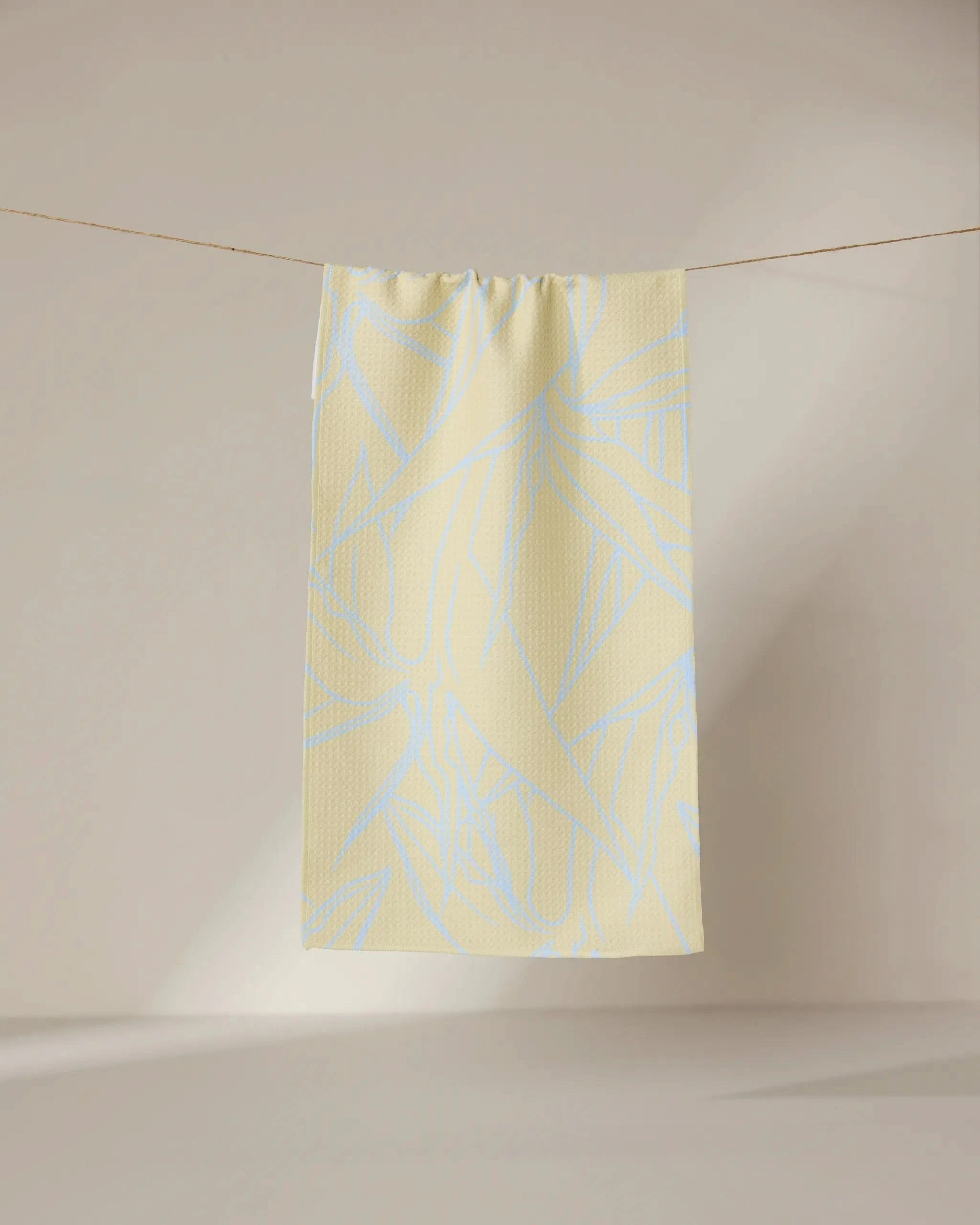 The GEOMETRY Paradise Blooms towel, featuring a pale blue abstract leaf design on light yellow, hangs neatly on a thin clothesline against a softly lit, neutral background.