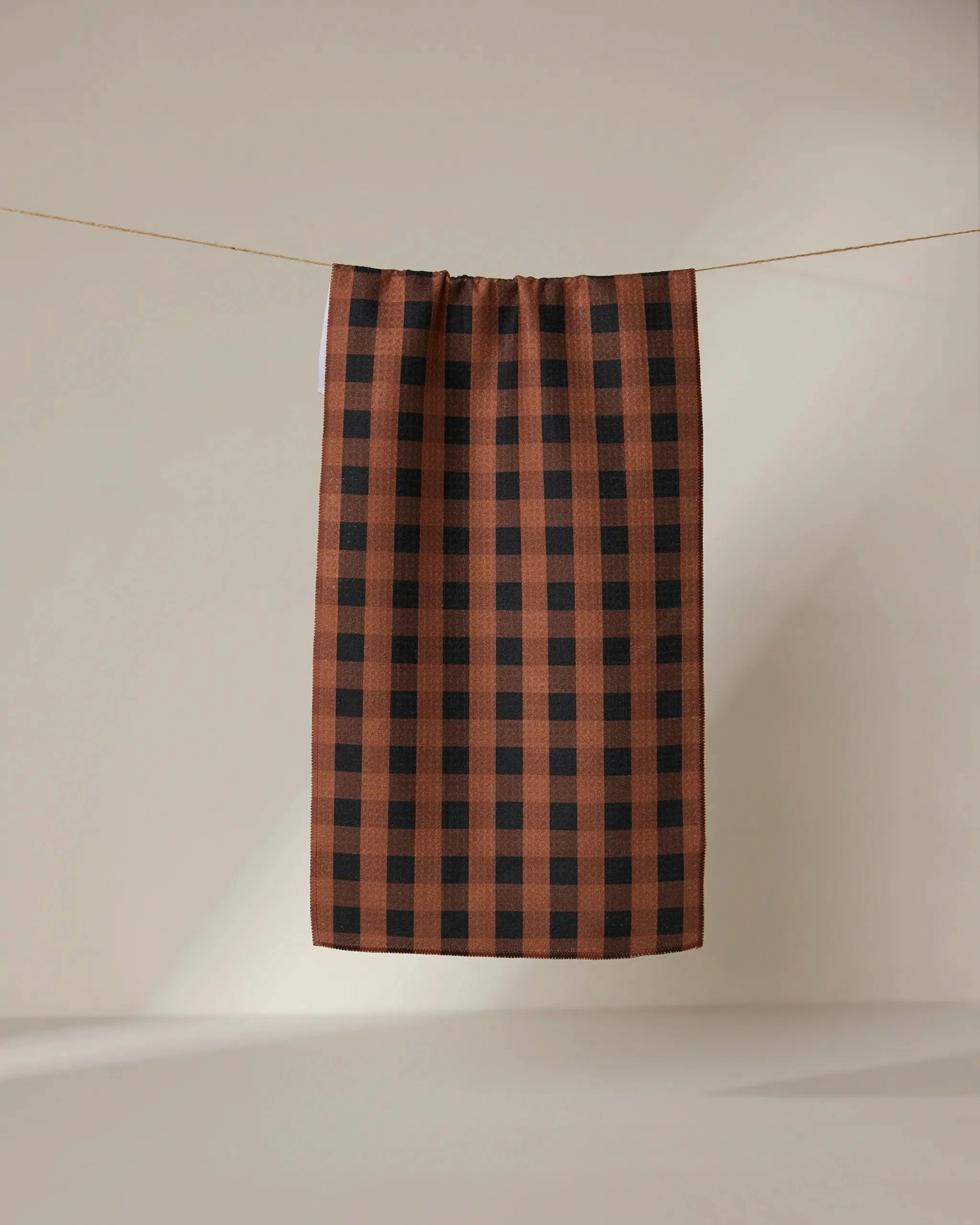 The GEOMETRY Fall Gingham towel, featuring a brown and black checkered pattern, hangs on a thin string against a neutral beige background.