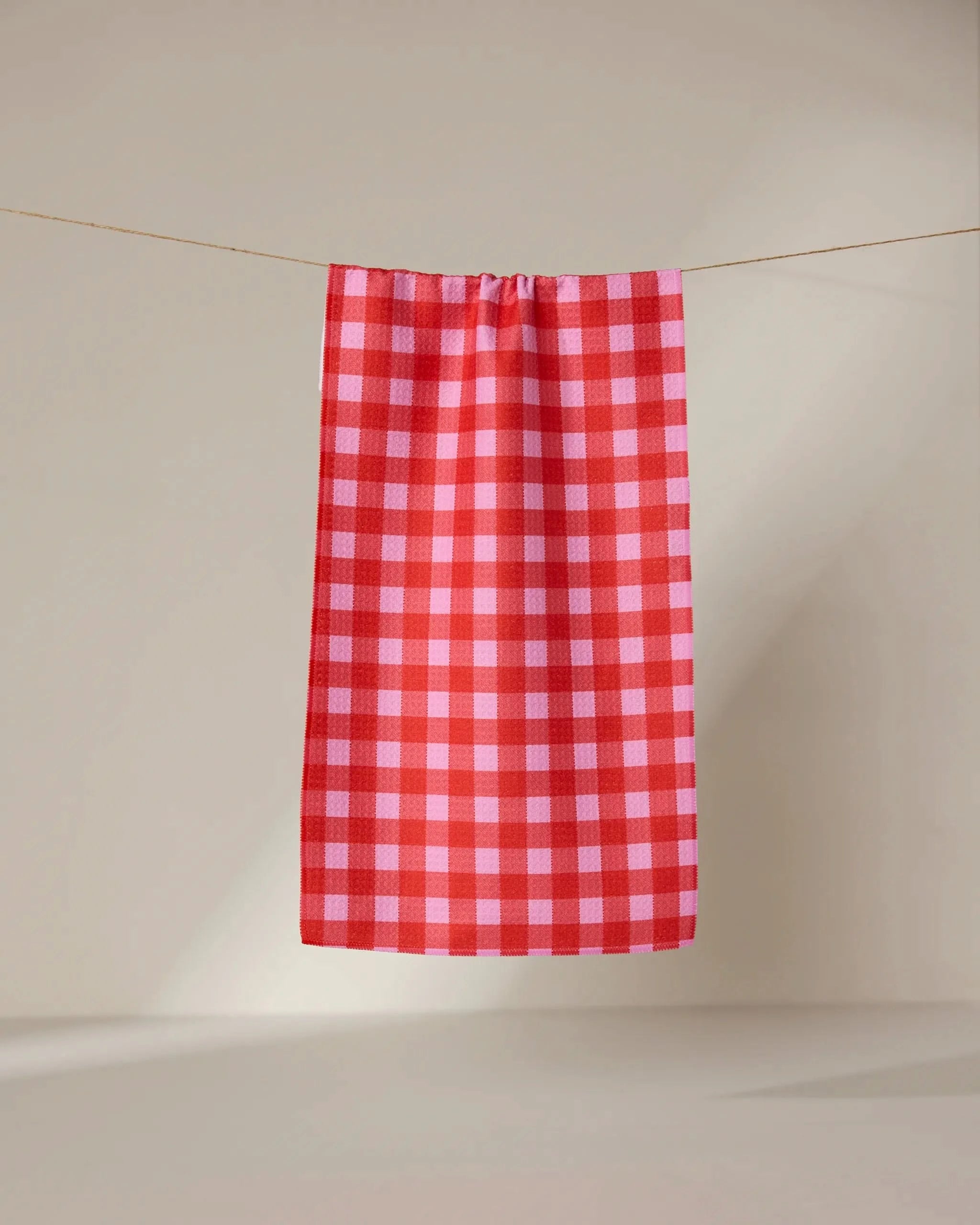 The GEOMETRY Cherry Gingham ultra-absorbent towel in red and pink checks hangs neatly on a clothesline against a plain, light background.