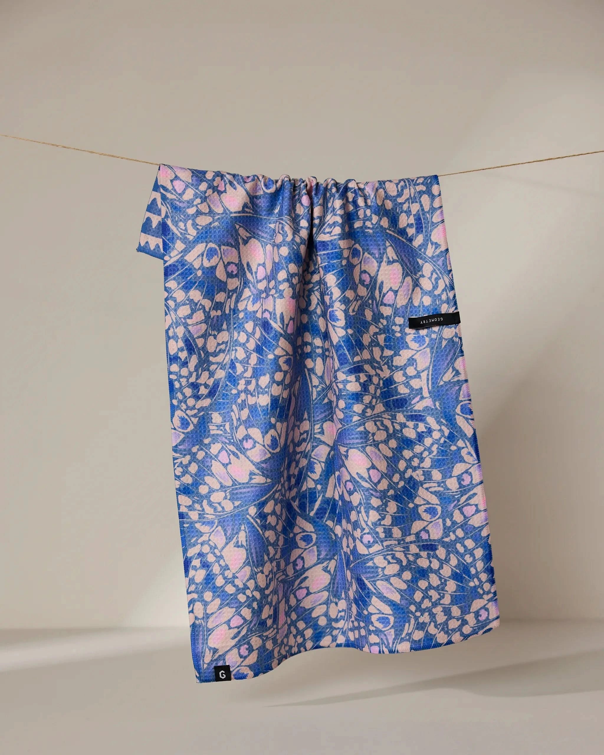 The Geometry Lav tea towel, featuring blue and pink abstract shapes and a small black tag, hangs on a clothesline against a plain light background. Its quick-drying fabric displays the brands signature modern pattern.