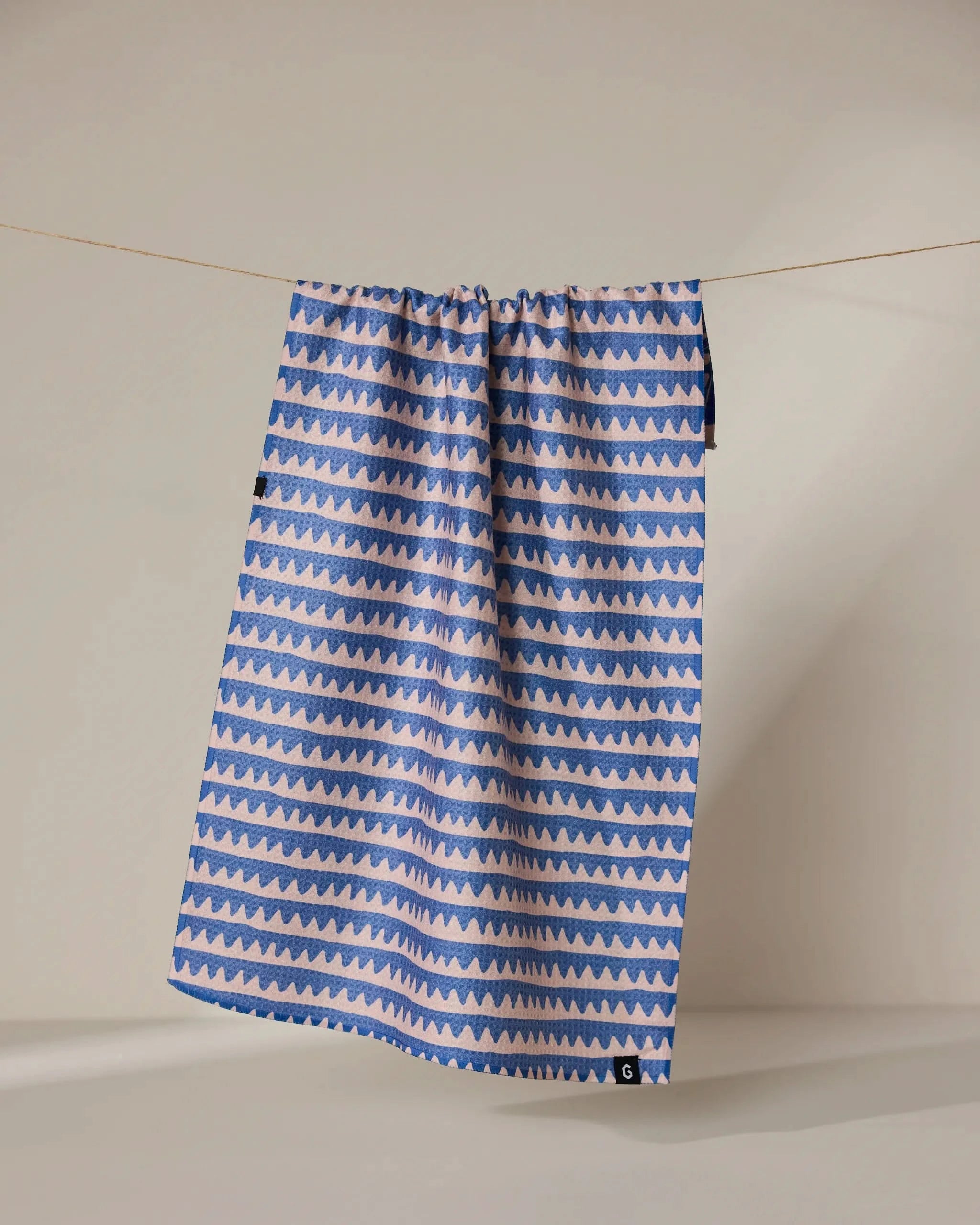 The Geometry Lav tea towel features double-sided alternating blue and light pink zigzag stripes, shown hanging on a clothesline against a neutral background.