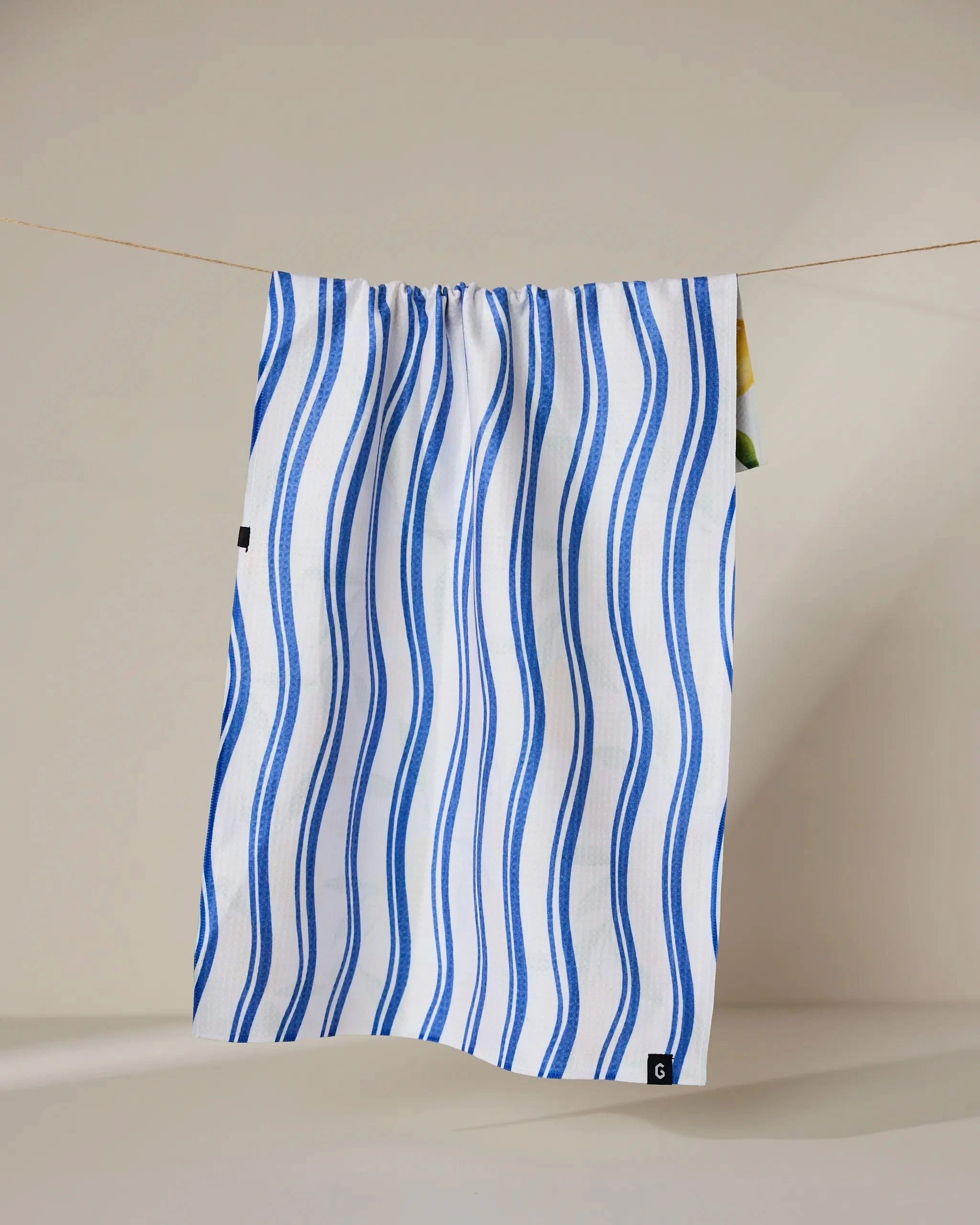 The Geometry Lemon Bliss tea towel, featuring wavy blue stripes on white fabric, hangs from a clothesline against a simple light background.