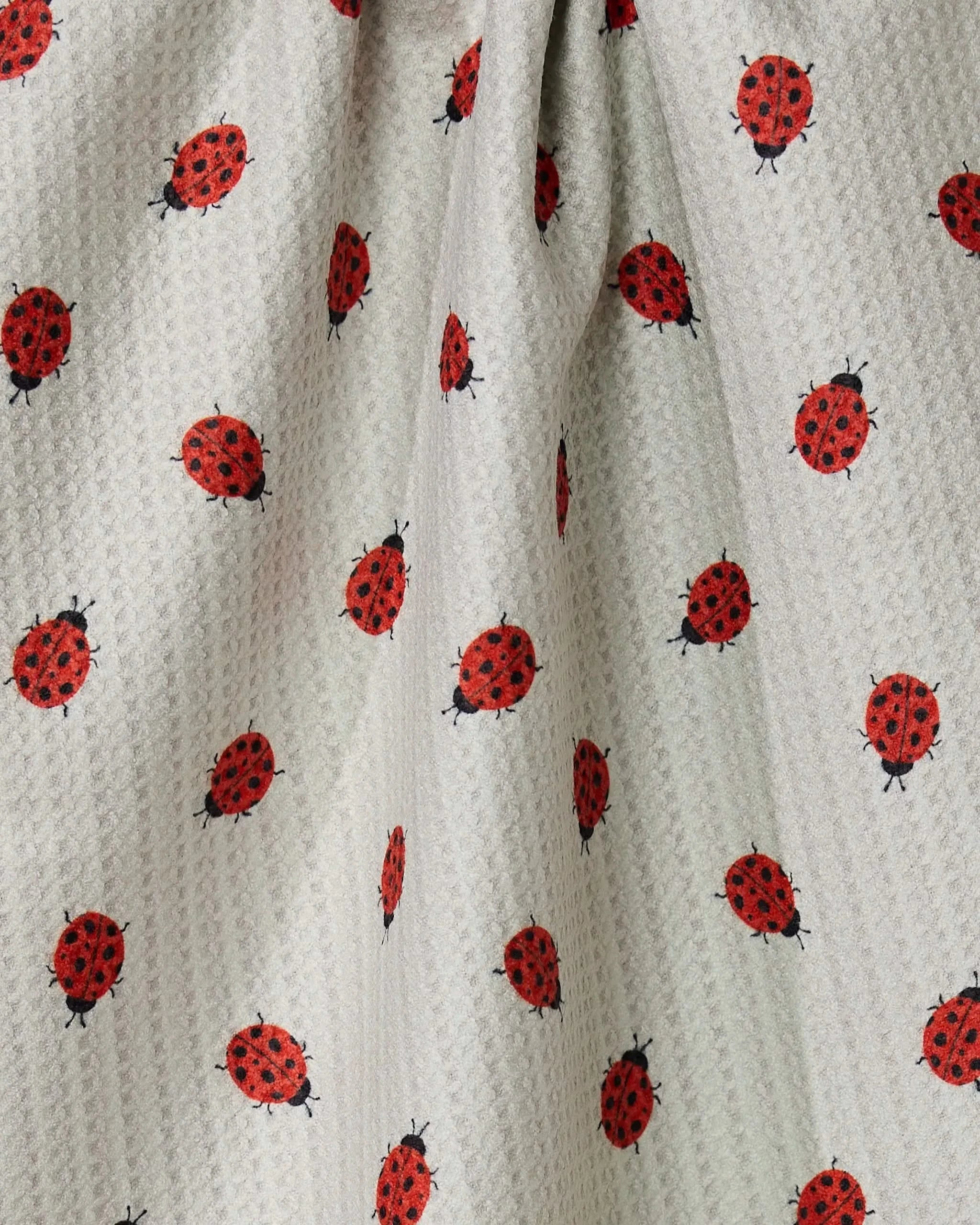 A close-up of the Geometry Lady tea towel, made from quick-drying, light-colored textured fabric with a repeating red and black ladybug pattern. The material is gently gathered, creating soft folds and shadows.