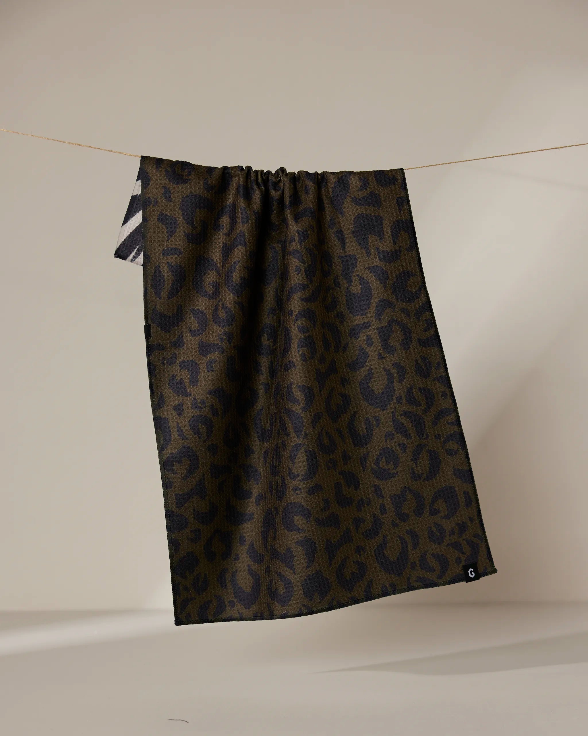 A dark brown and black leopard print Geometry Safari Absorbent Quick-Dry Tea Towel hangs on a thin clothesline against a neutral background, featuring a small white G in one corner and double-sided design.