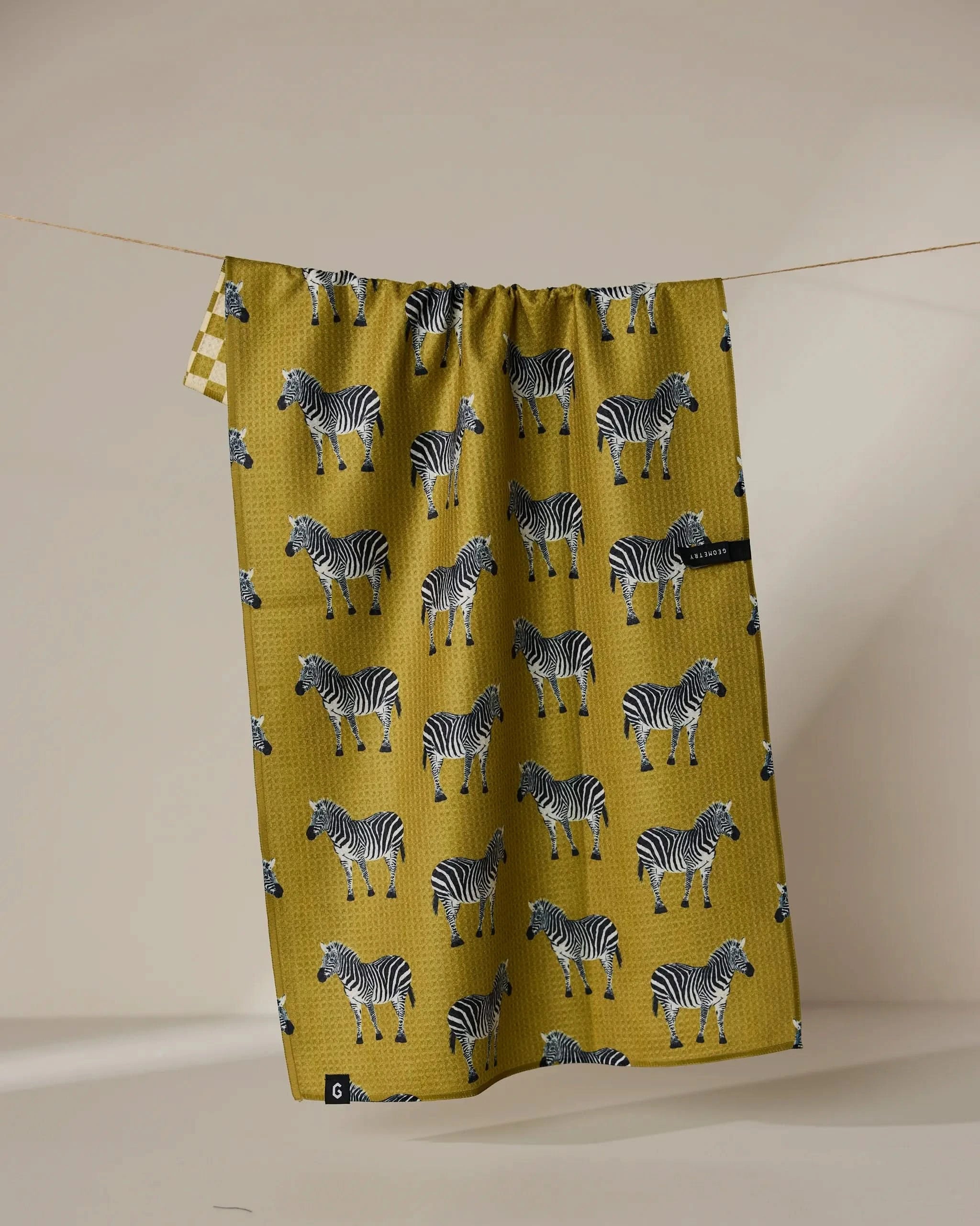 The Geometry Zebra Way tea towel, featuring a quick-drying fabric and repeating zebra pattern in mustard yellow, hangs on a string against a plain beige background.