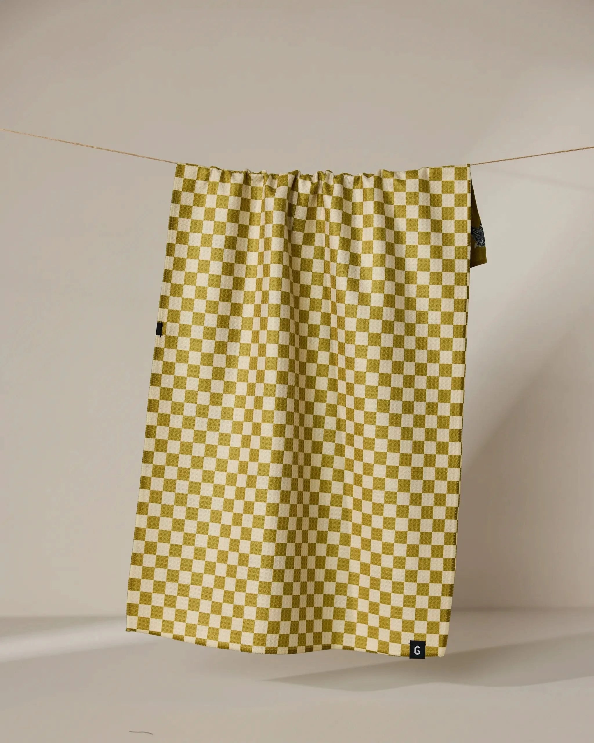 The Geometry Zebra Way tea towel in mustard yellow and white check pattern hangs from a thin string against a beige background, with a small black tag on its lower right corner.