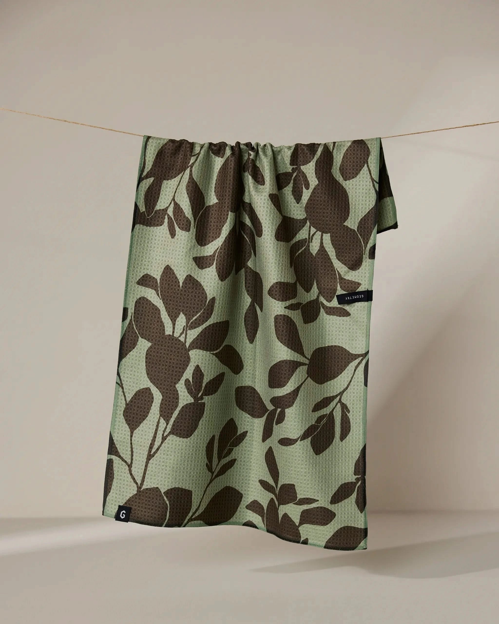 The Geometry Eucalyptus towel, featuring large dark brown leafy patterns on green and a small black tag at the edge, hangs on a string against a neutral background.